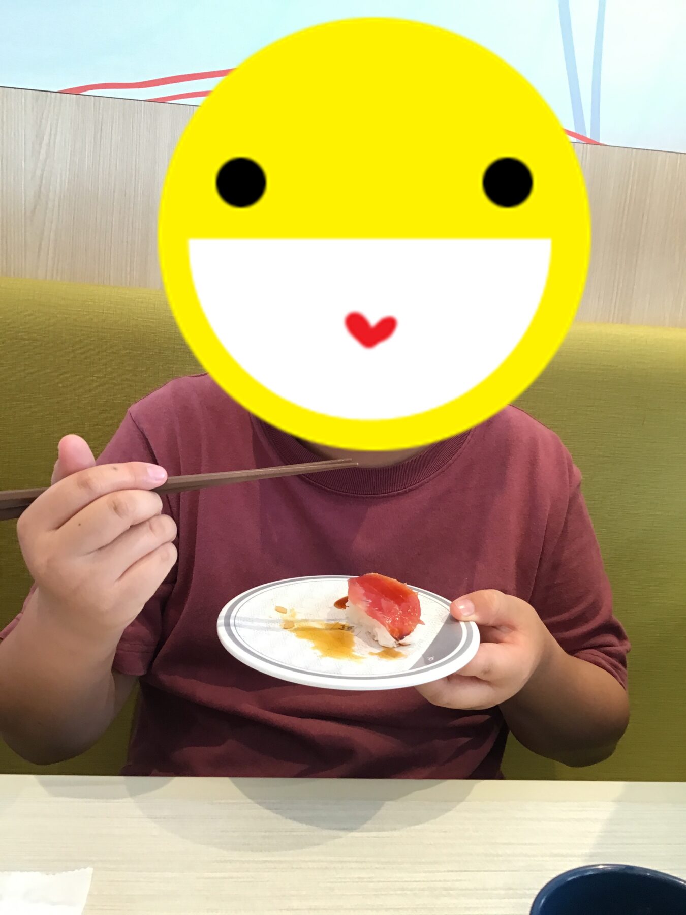 Person in a maroon shirt uses chopsticks to eat sushi at a restaurant, with a large yellow smiley emoji covering their face.