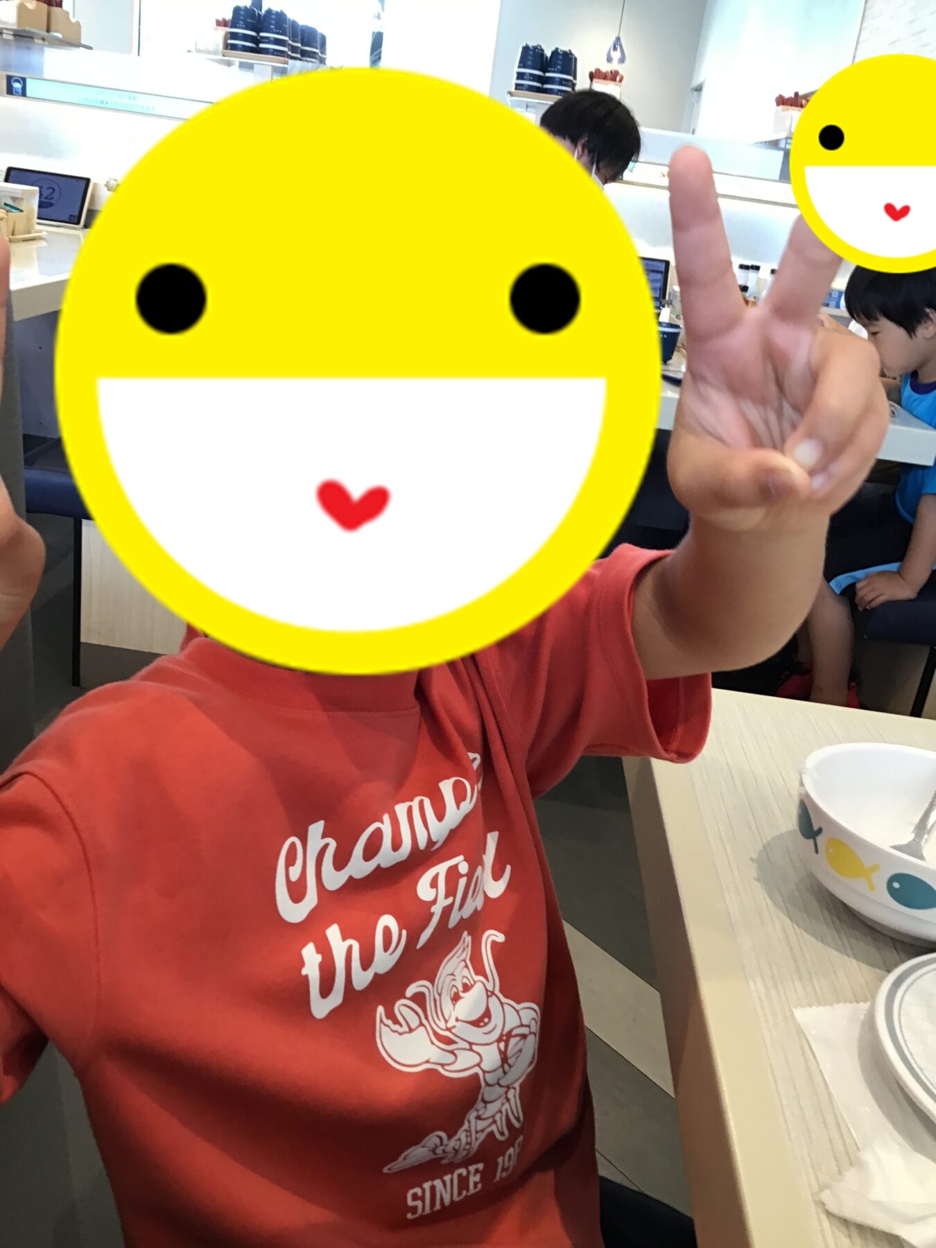 Child in a red shirt sits at a cafe, flashing a peace sign; a large yellow emoji graphic covers their face for privacy.