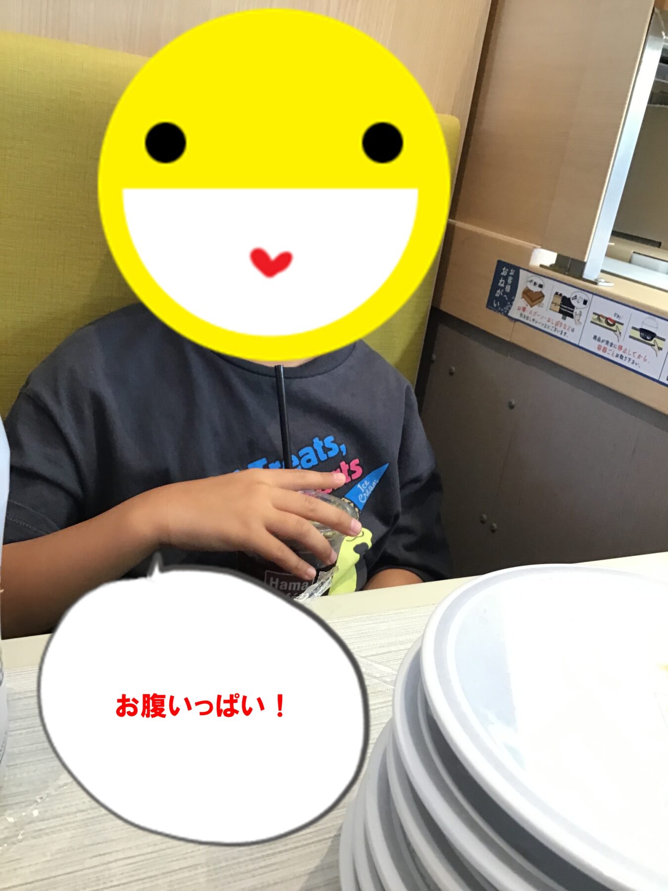 Person seated at a table in a restaurant with a large yellow smiley covering their face; speech bubble reads 'お腹いっぱい！' (I'm full!).