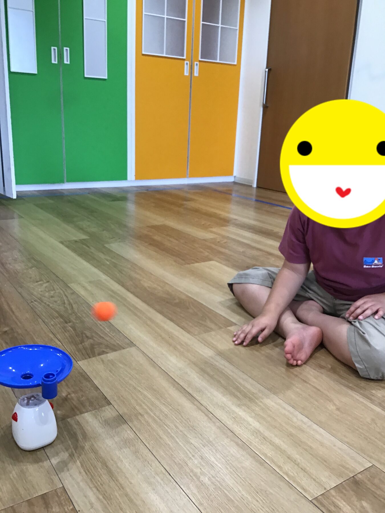 Child sitting cross‑legged on a wooden floor indoors, playing with a small orange ball; blue toy in the foreground and green/orange storage cabinets in the background.