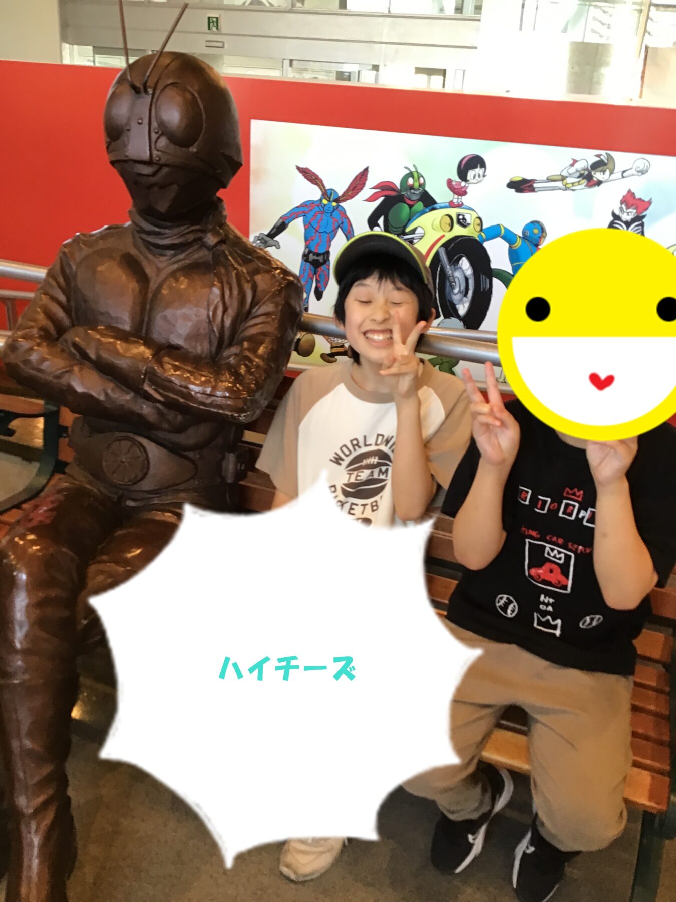 Two kids posing with a bronze armored statue indoors, one making a peace sign with a cheerful smile, background shows a colorful cartoon poster.