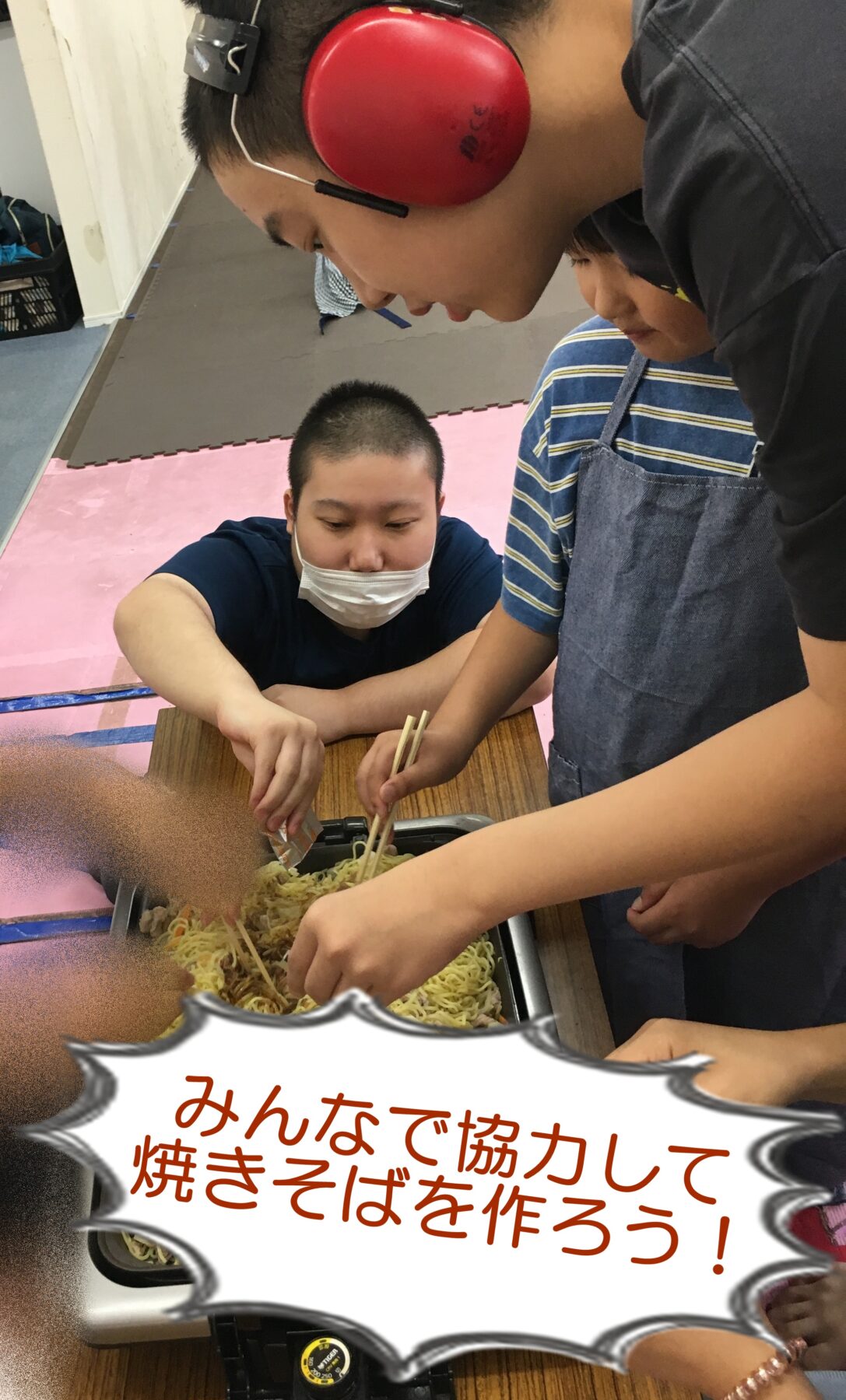 People cooking stir-fried noodles together at a table; one person wearing red earmuffs and a mask guides the others while using chopsticks.