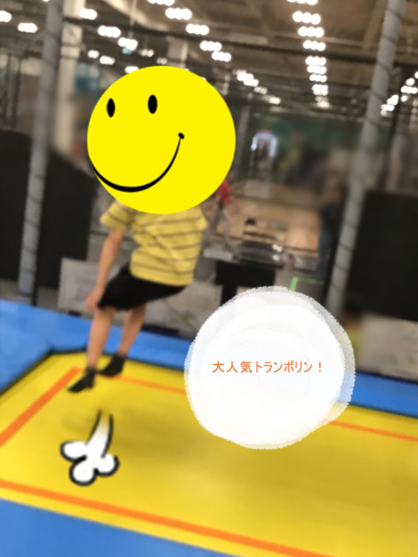 Child in a yellow-striped shirt stands on a bright yellow trampoline inside an indoor park, face covered by a large yellow smiley sticker.