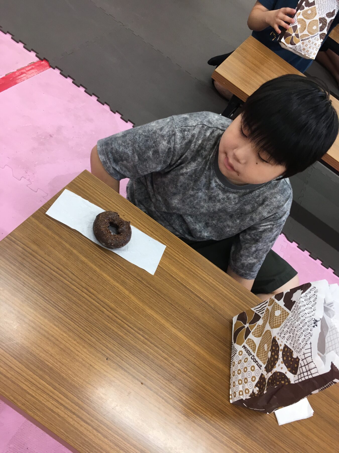 Young boy sitting at a wooden table with a chocolate donut on a napkin in a playroom area with pink foam mats nearby