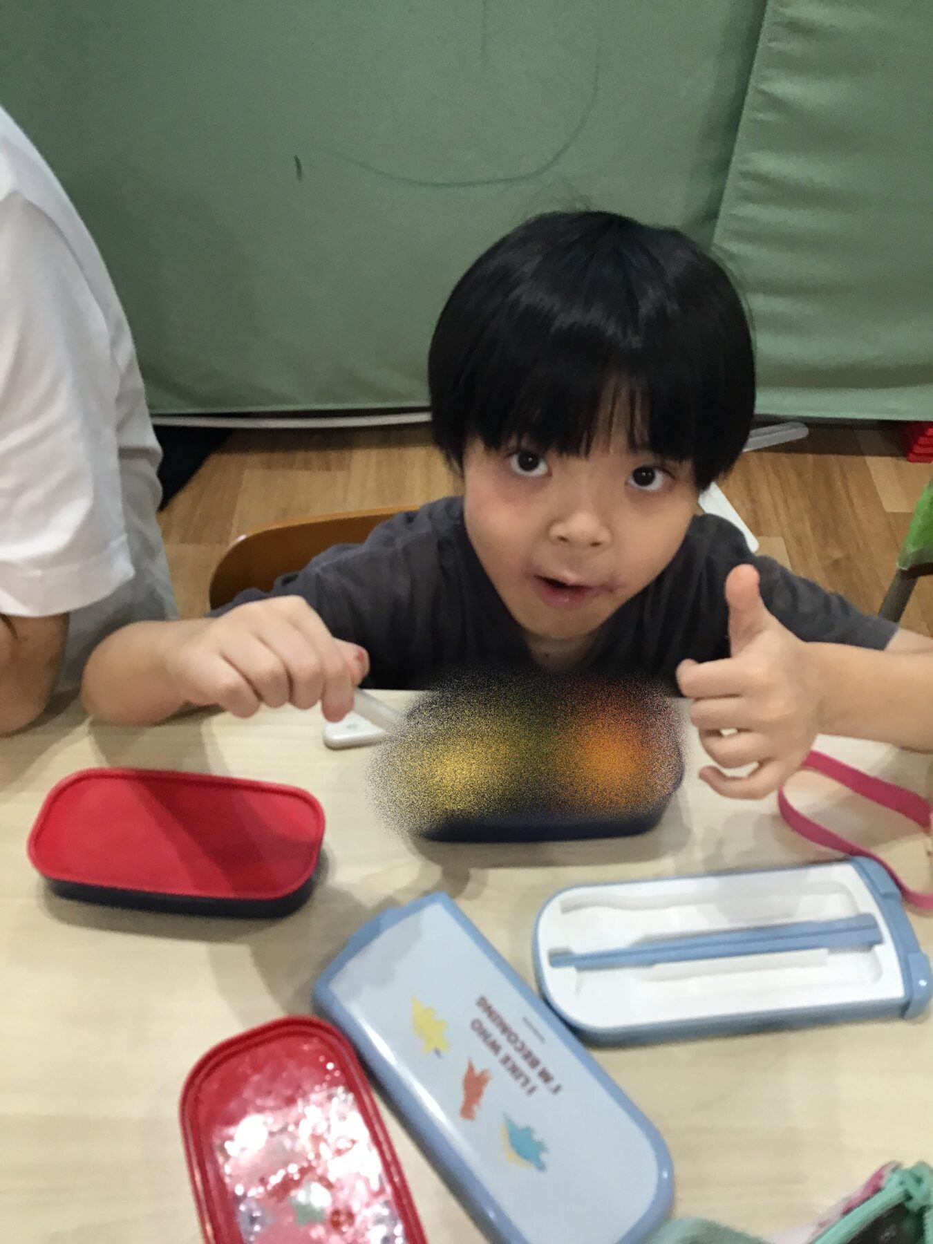 Young boy with black hair sits at a table, giving a thumbs-up with lunch boxes and utensils in front.