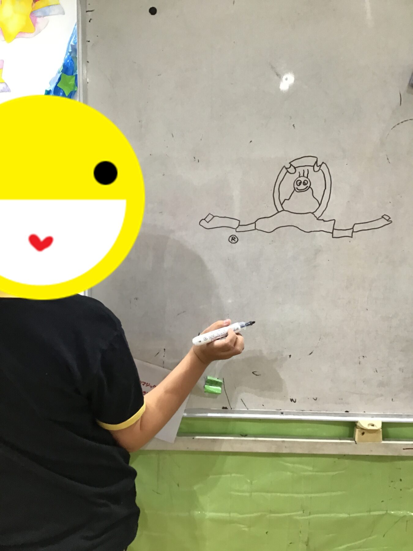 Person wearing a black shirt draws a cartoon figure on a gray wall with a marker; a large yellow emoji sticker covers the left side in the foreground.