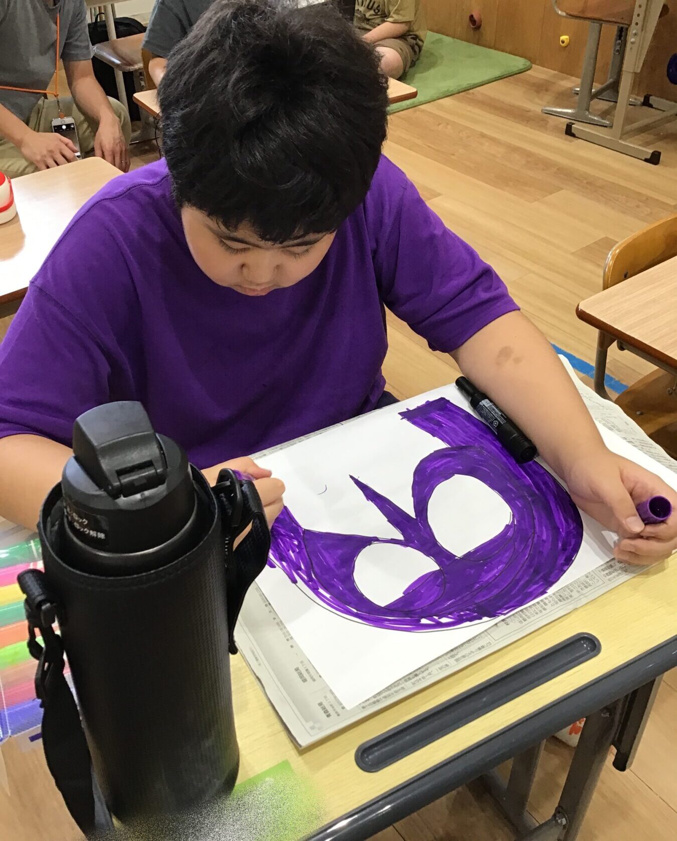 Child in a purple shirt drawing with a purple marker at a desk, artwork in progress; a black water bottle is in the foreground on the desk in a classroom setting.