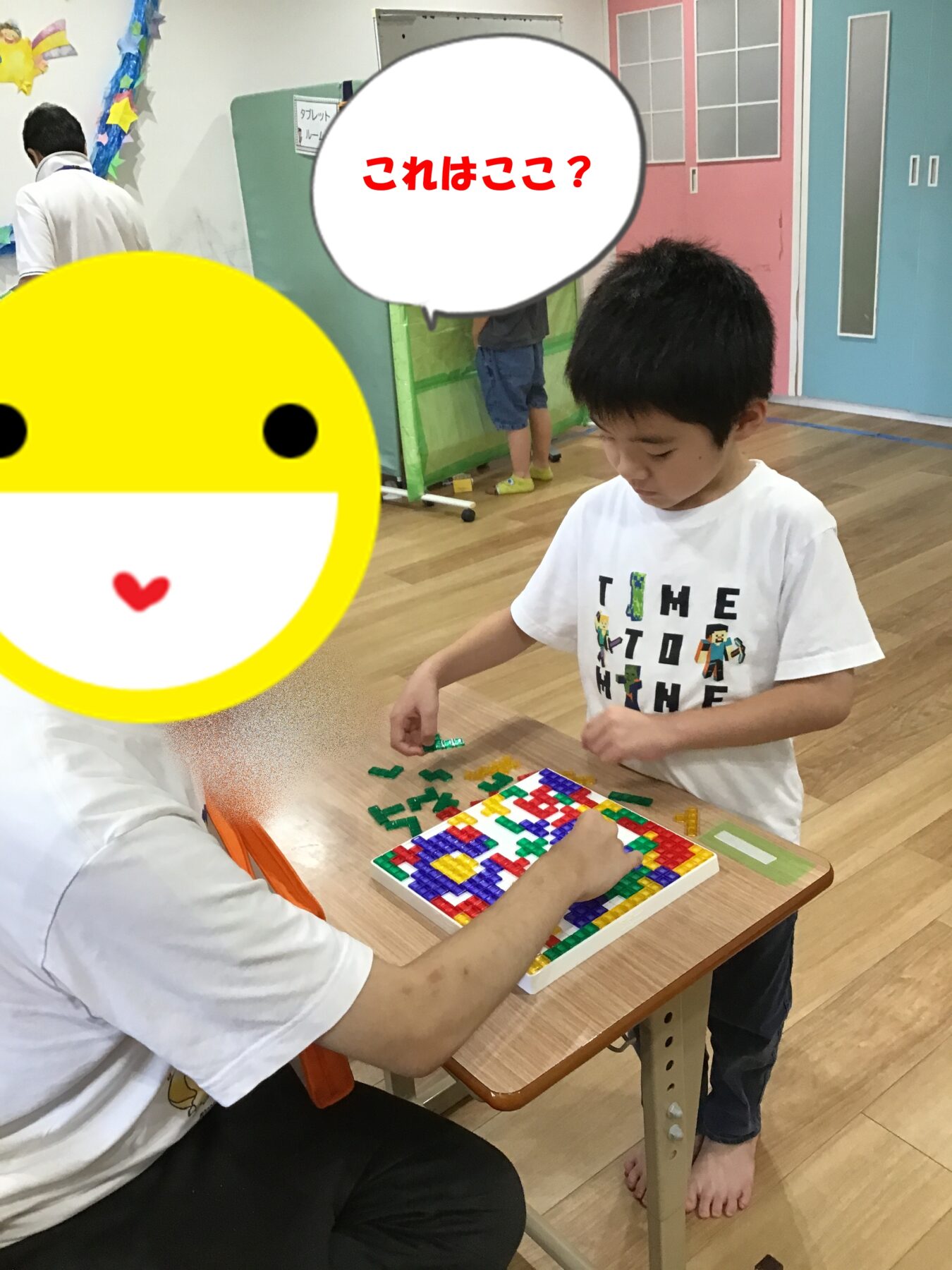 Child stands at a table placing colorful puzzle tiles while an adult sits nearby, guiding the activity; a large yellow emoji obscures part of the scene.