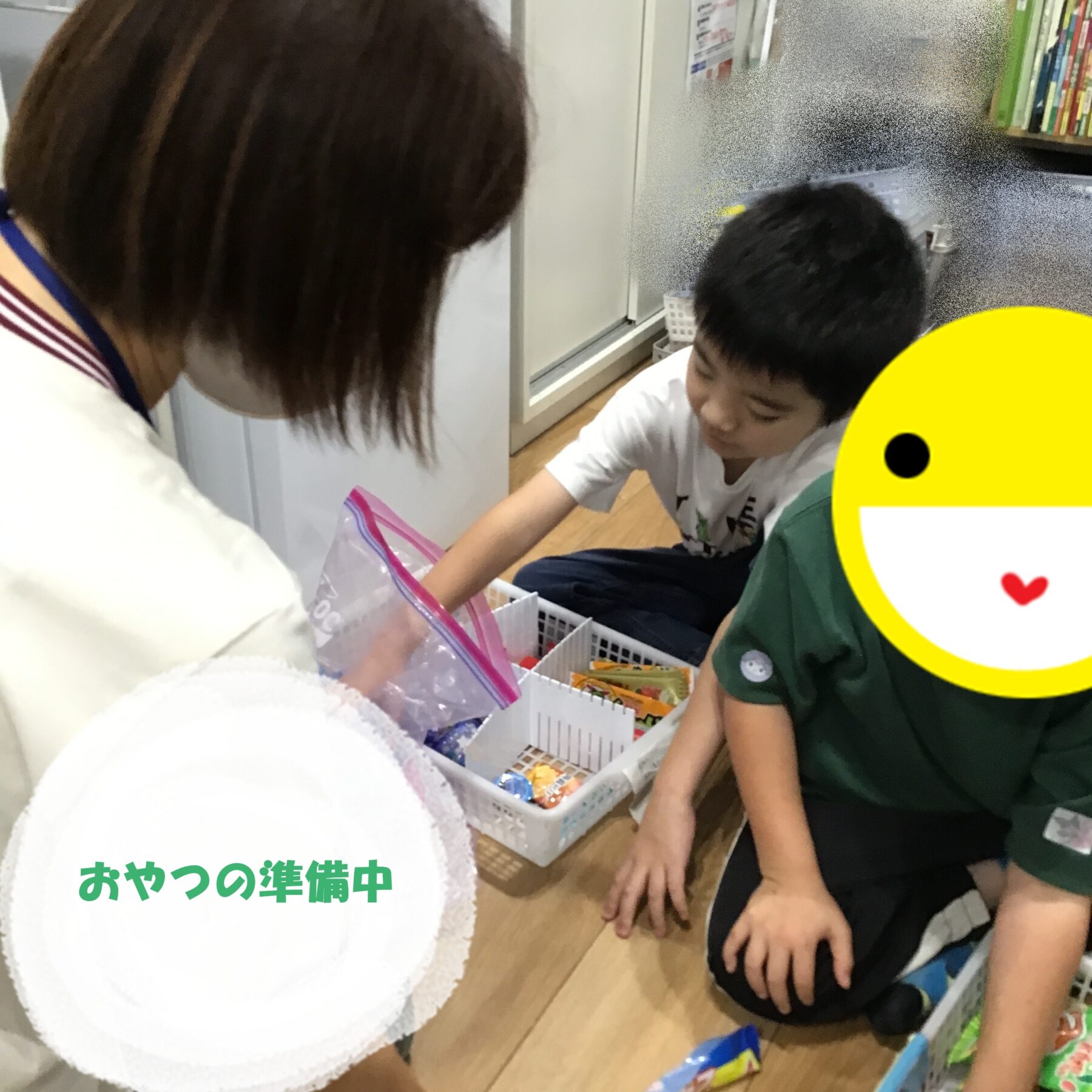 Two children kneel on the floor, sorting snack bags into white baskets in a store, with a large yellow emoji sticker on the right.