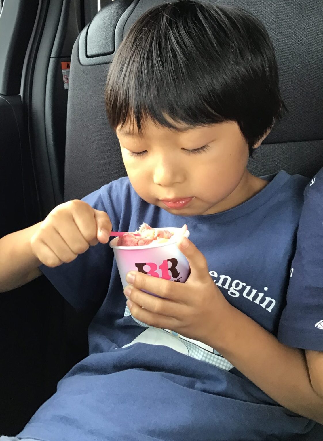 Young child with dark hair in a blue shirt, seated in a car, eating pink ice cream from a small cup with a pink spoon.