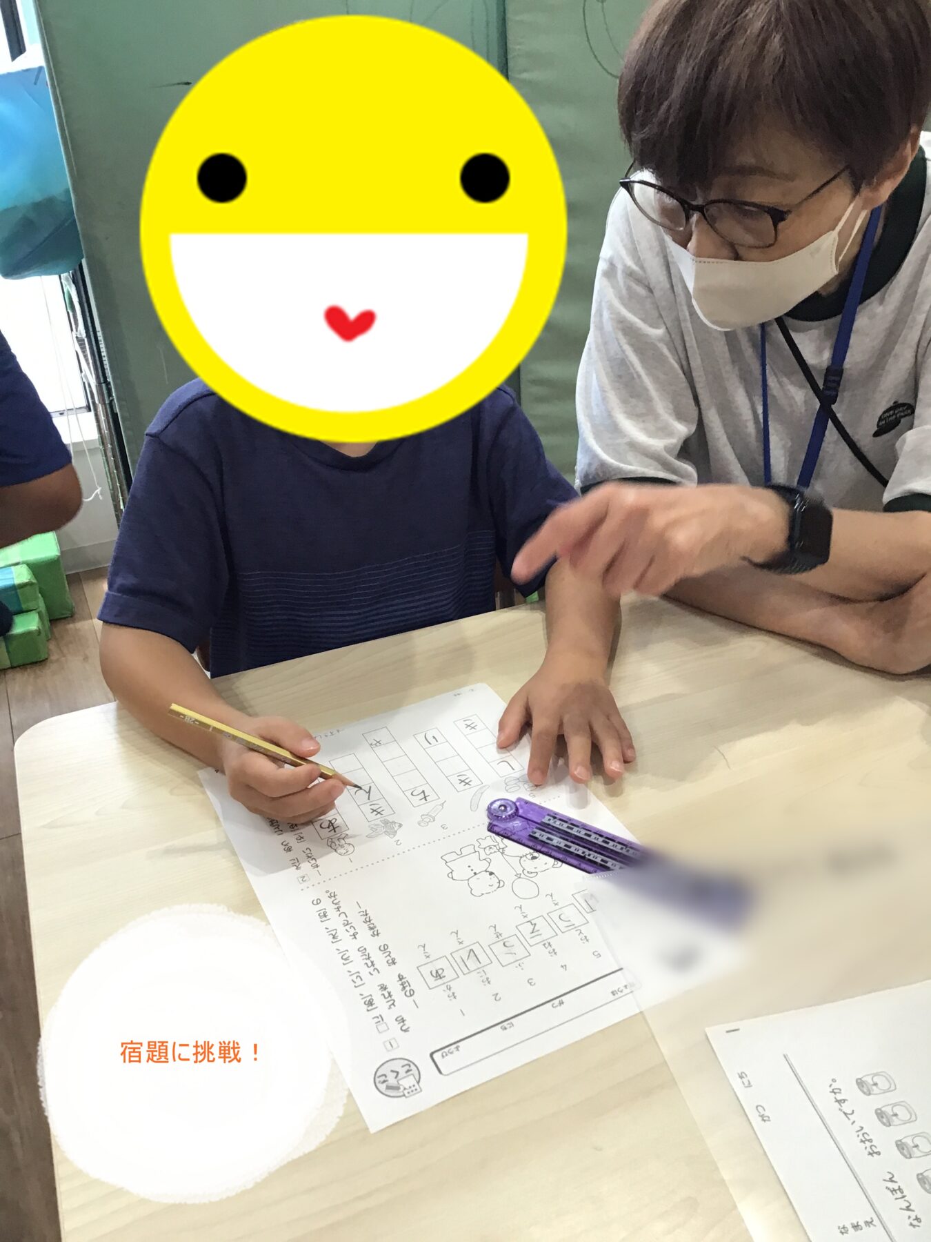 Child in a blue shirt writes on a worksheet at a table while an adult wearing glasses and a mask guides him.