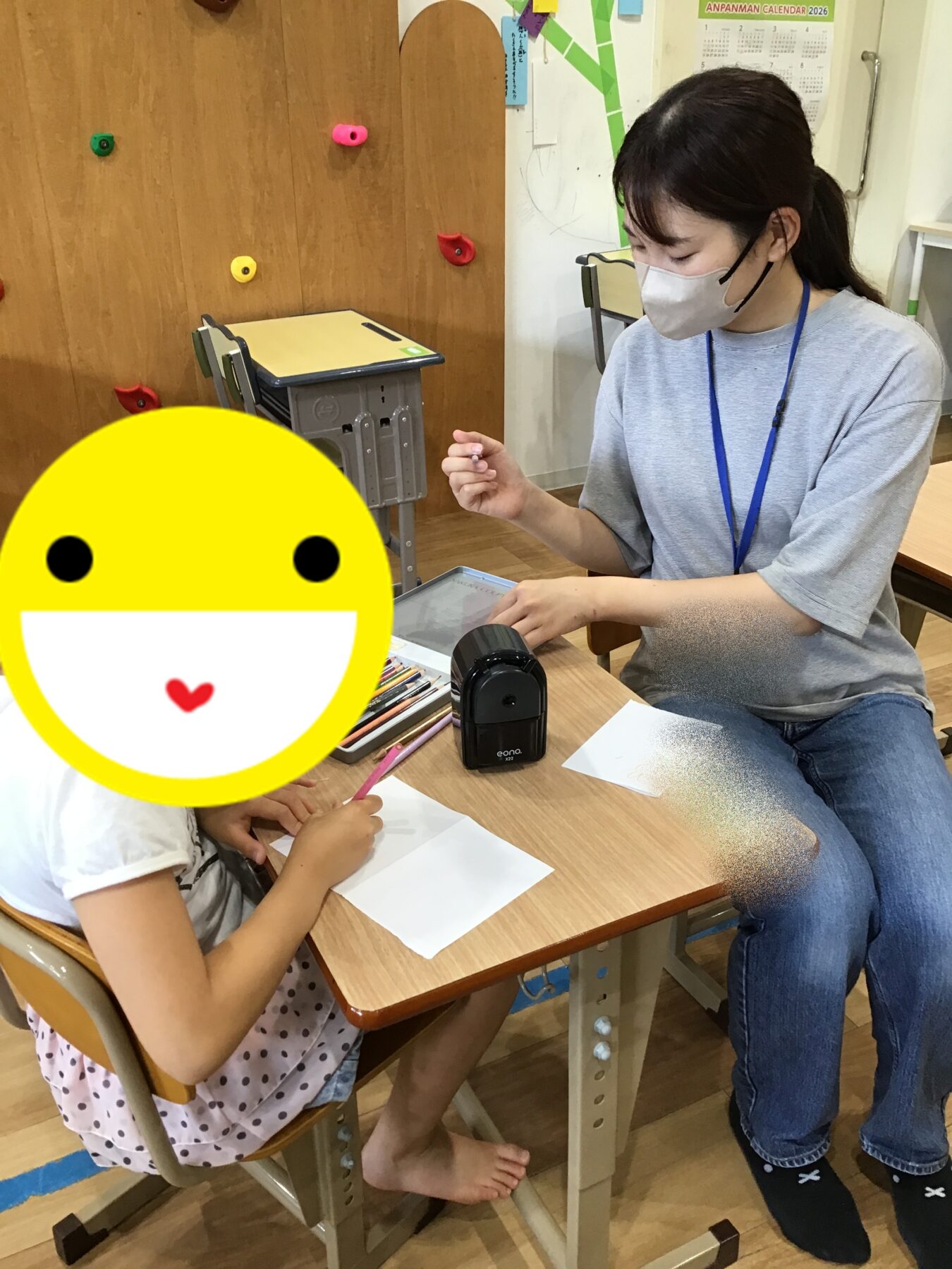 Teacher wearing a mask and blue lanyard helps a student at a desk with drawings in a classroom; pencil sharpener and colored pencils nearby.