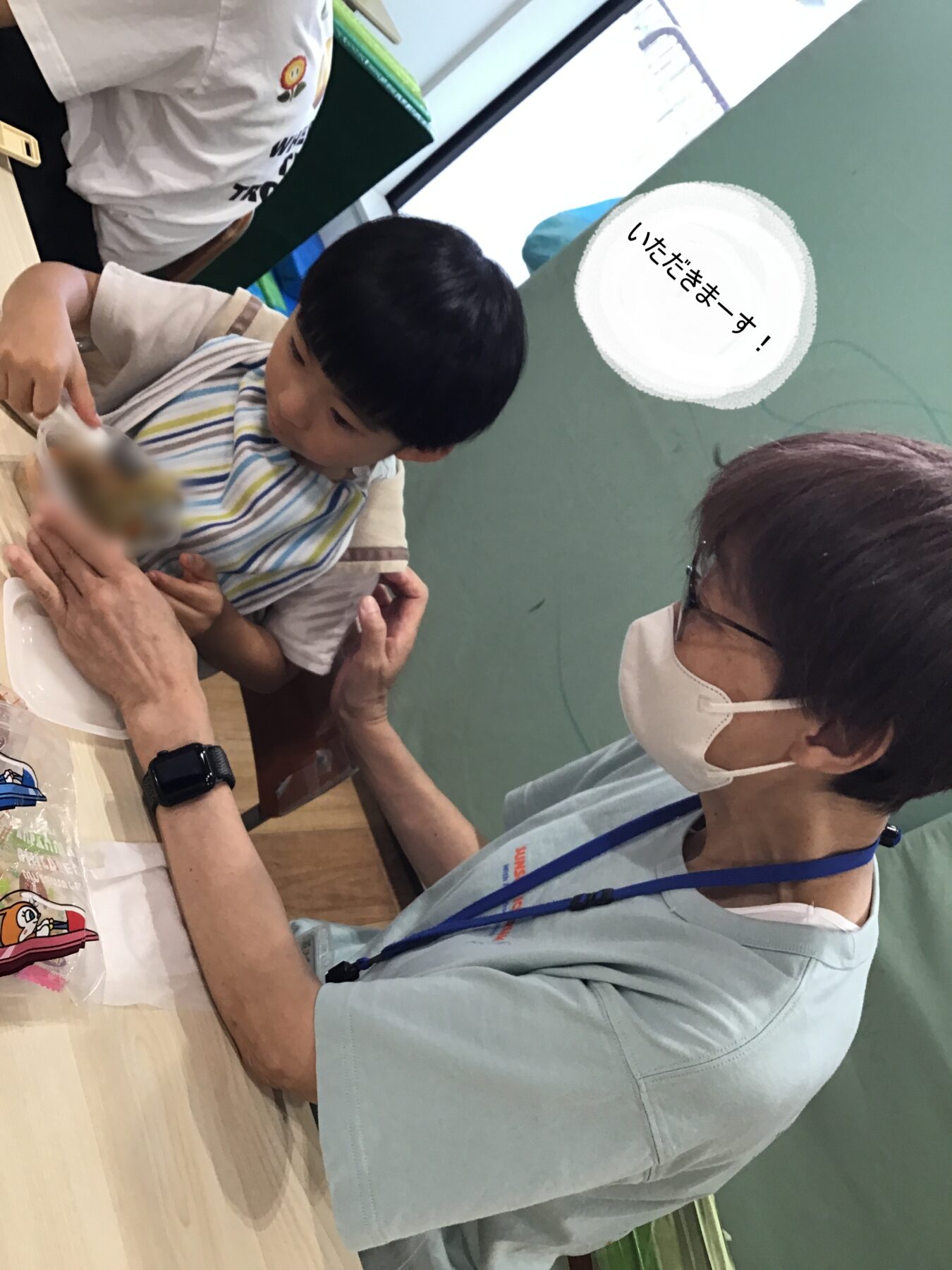 Child in a striped bib being fed by an adult at a table; another adult in a mask nearby; speech bubble reads 「いただきます!」.