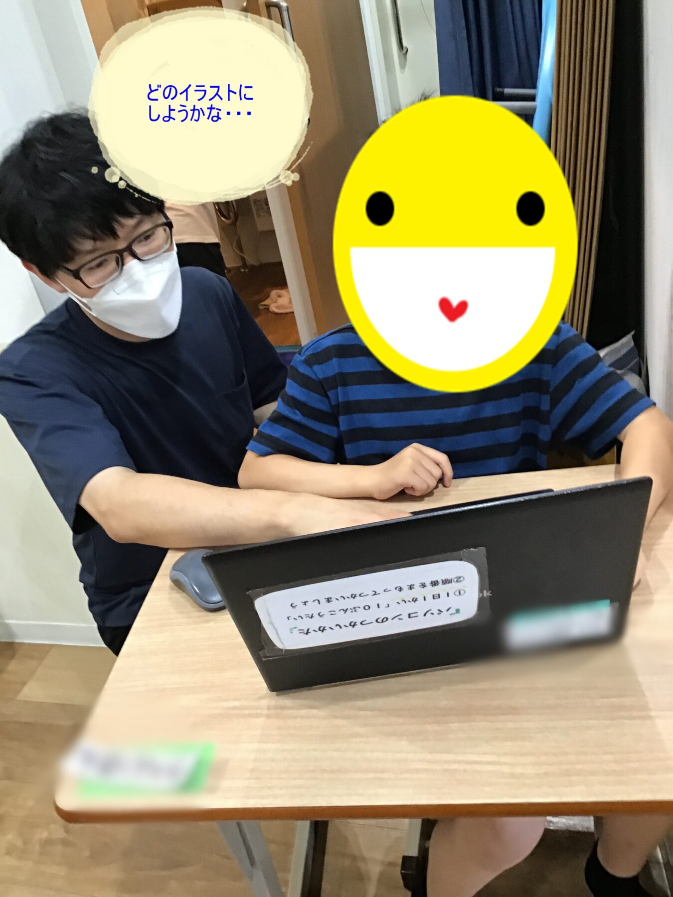 Adult tutor in a mask helps a child sit at a desk with a laptop; the child's face is covered by a yellow smiley sticker.