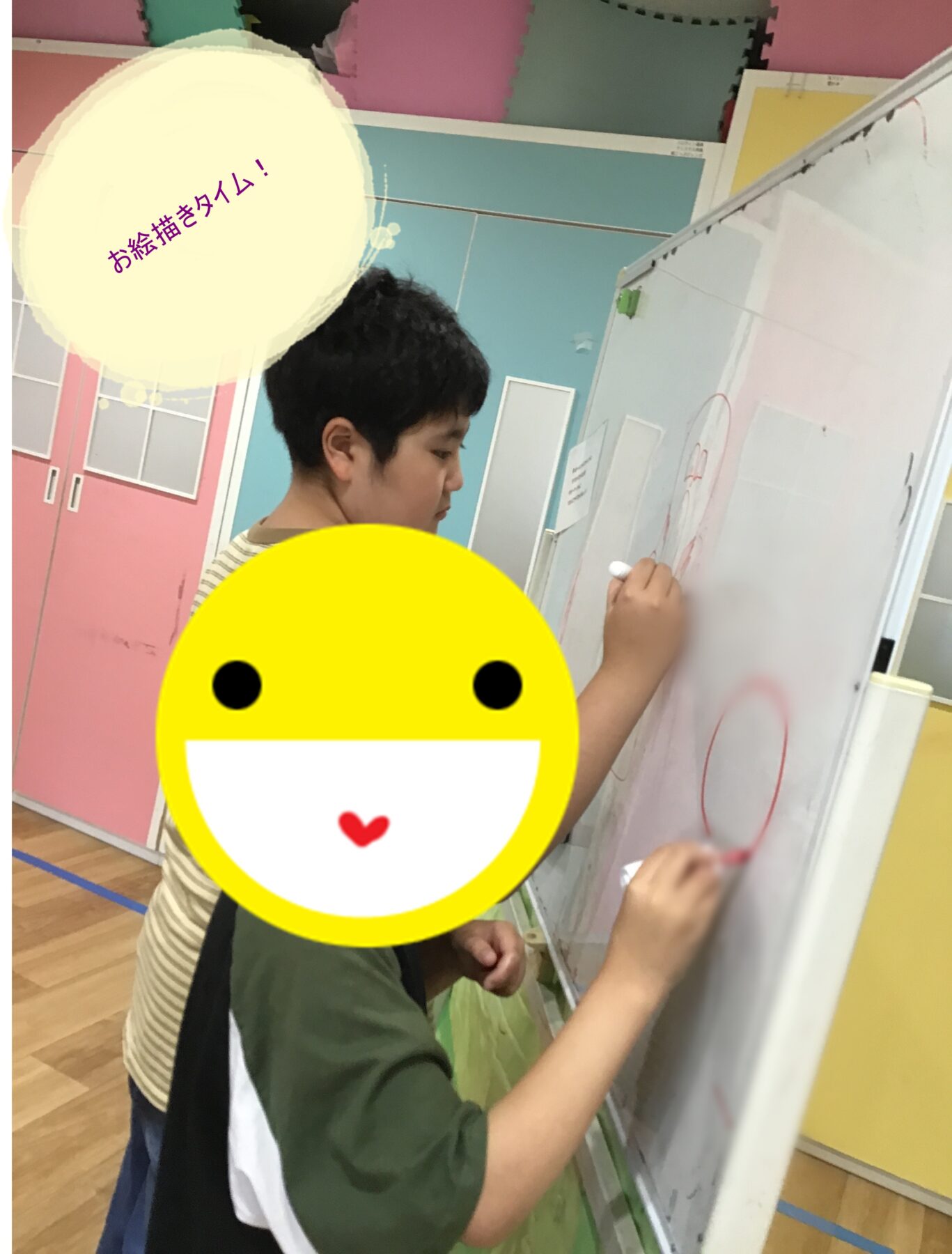 Child wearing a striped shirt and green sleeves draws on a whiteboard in a bright, colorful classroom, using a marker with another student nearby.