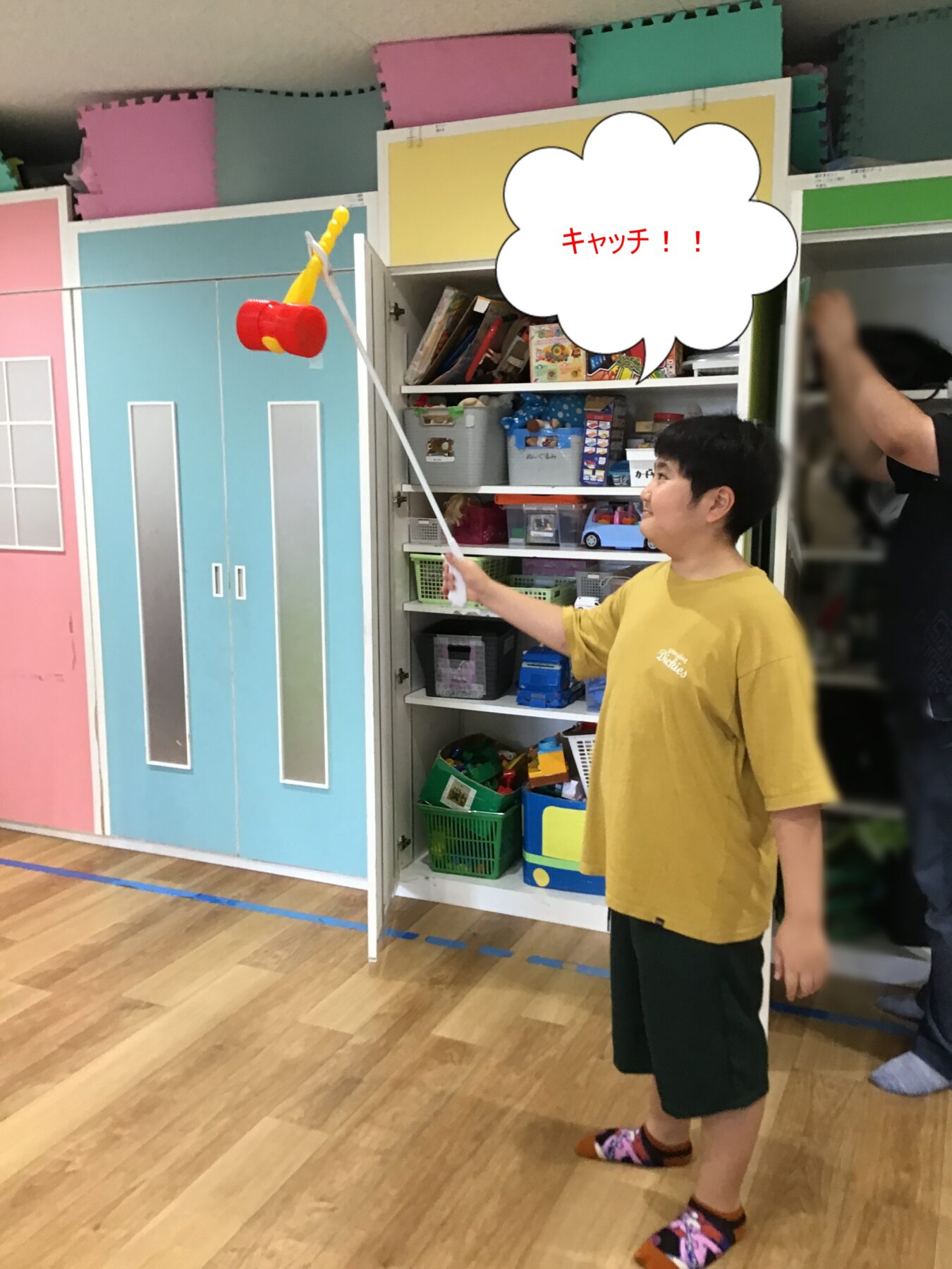 Boy in a yellow shirt holds a long pole with a red toy hammer, standing in a colorful playroom with shelves of toys in the background.