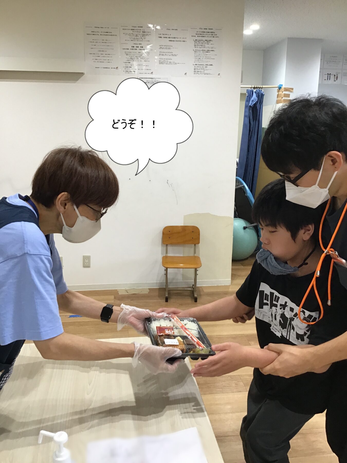 Healthcare worker wearing a mask and gloves hands a packaged meal to a man also wearing a mask, across a table in a room.