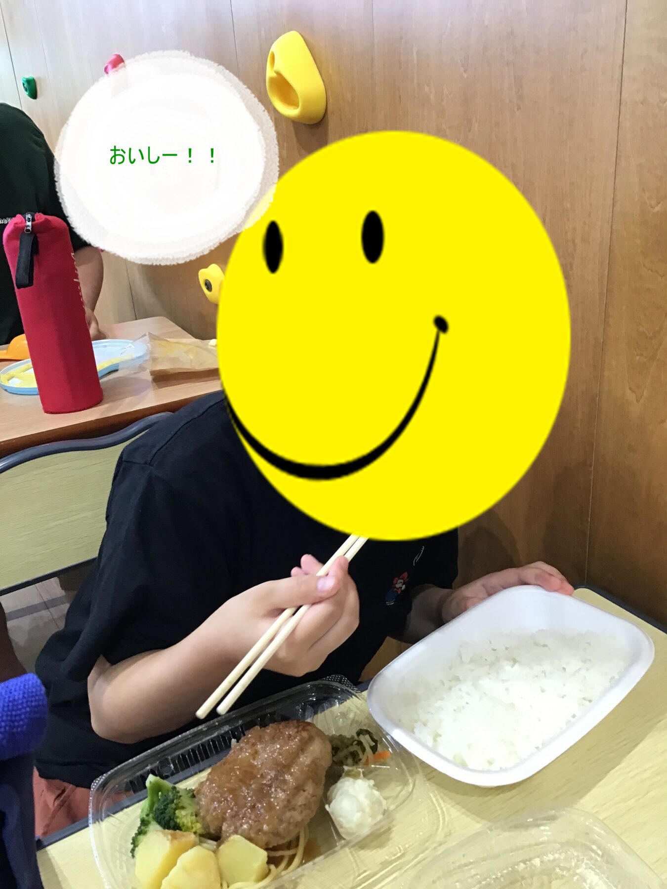 Child eating with chopsticks at a table; bento-style meal with rice, potatoes, broccoli, and meat; yellow smiley face overlayed on head.