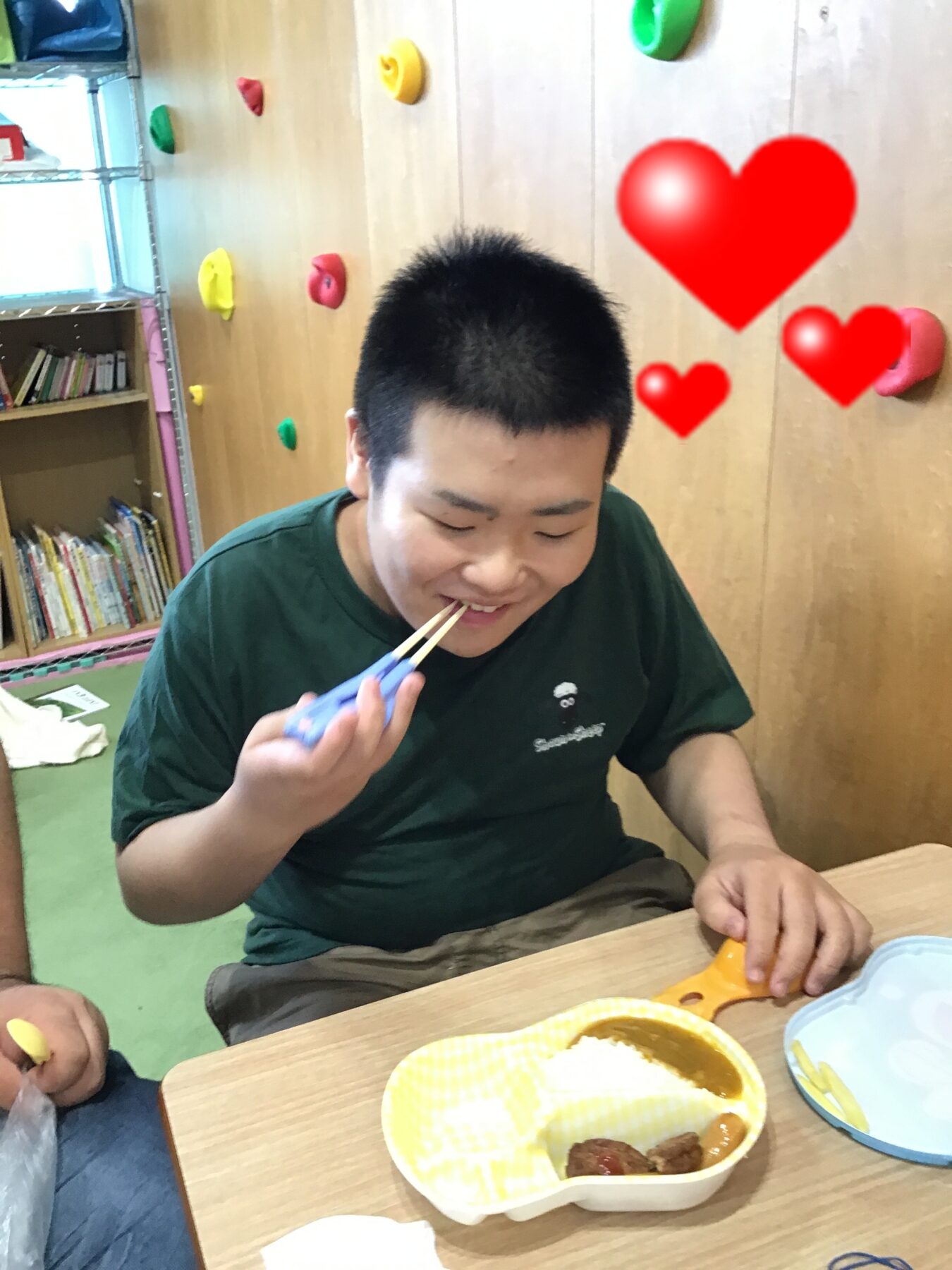 Person in a green shirt uses chopsticks to eat from a yellow-patterned container at a wooden table in a playroom or classroom, with climbing holds on the wall in the background and red heart overlays nearby.