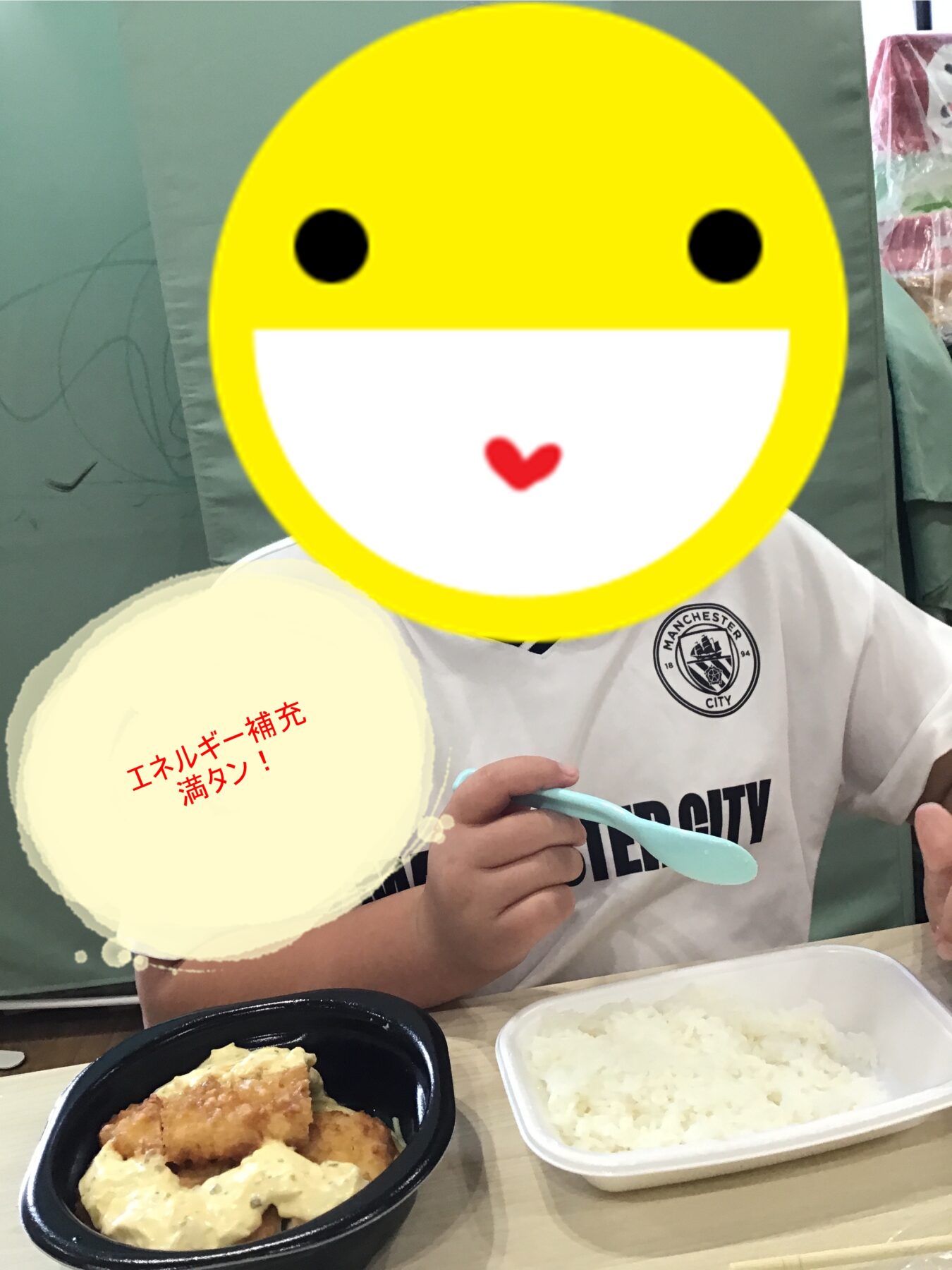 Person wearing a Manchester City jersey sits at a table; their face is covered by a large yellow smiley sticker while they hold a spoon with rice and fried food in front of them.
