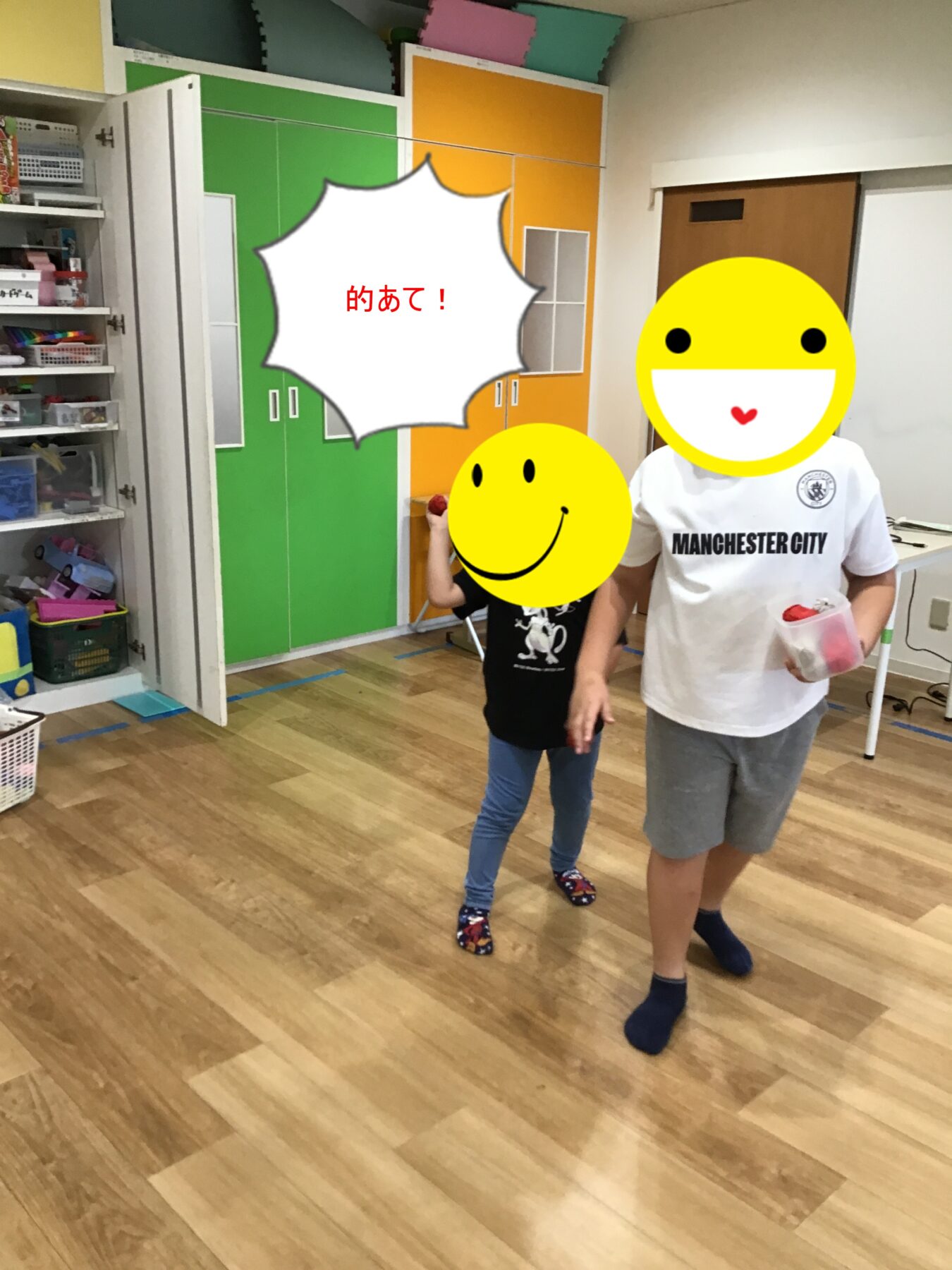 Two children stand indoors in a classroom or playroom, smiling emoji overlays cover their faces while one holds a small bag of items; colorful green and orange closet panels are in the background. A speech bubble with red Japanese text is centered in the scene.