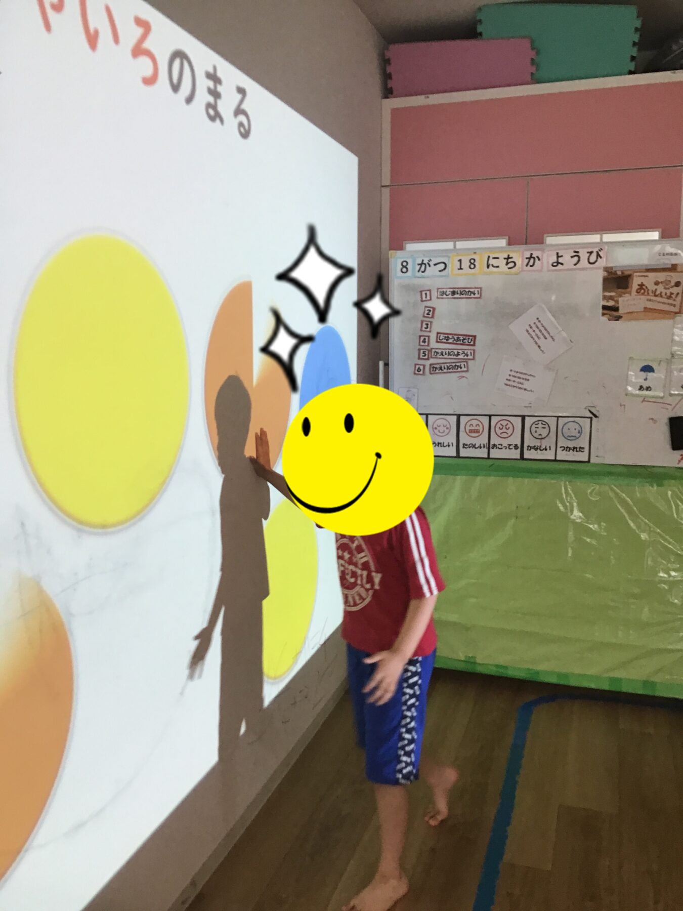 Child in red shirt and blue shorts stands barefoot, painting a mural with large yellow ovals on a wall, smiley sticker covering face.