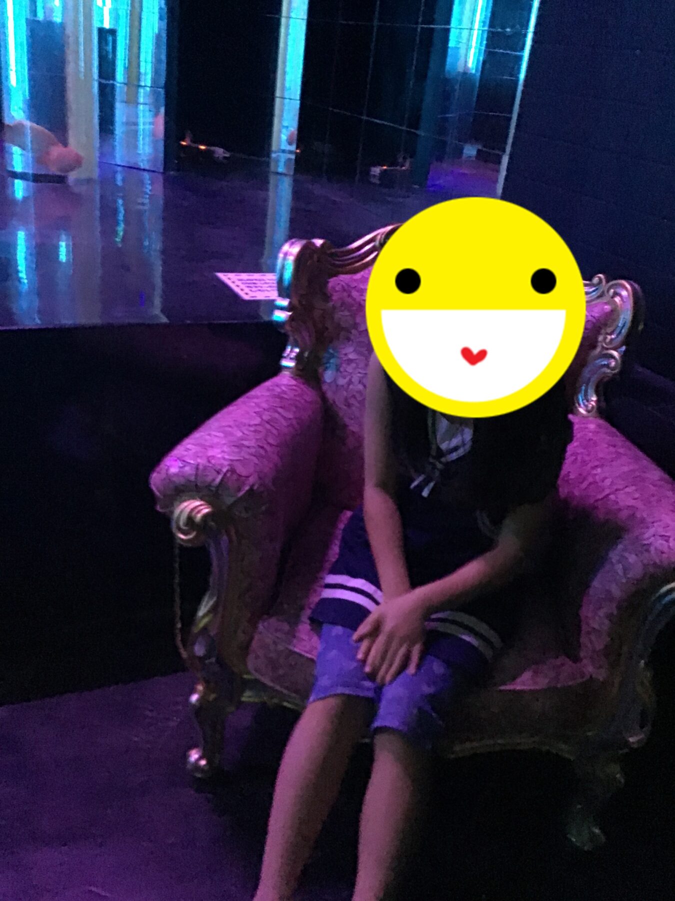 Two people sit on a pink ornate sofa in a neon-lit, dark room; faces covered by a yellow smiling emoji.
