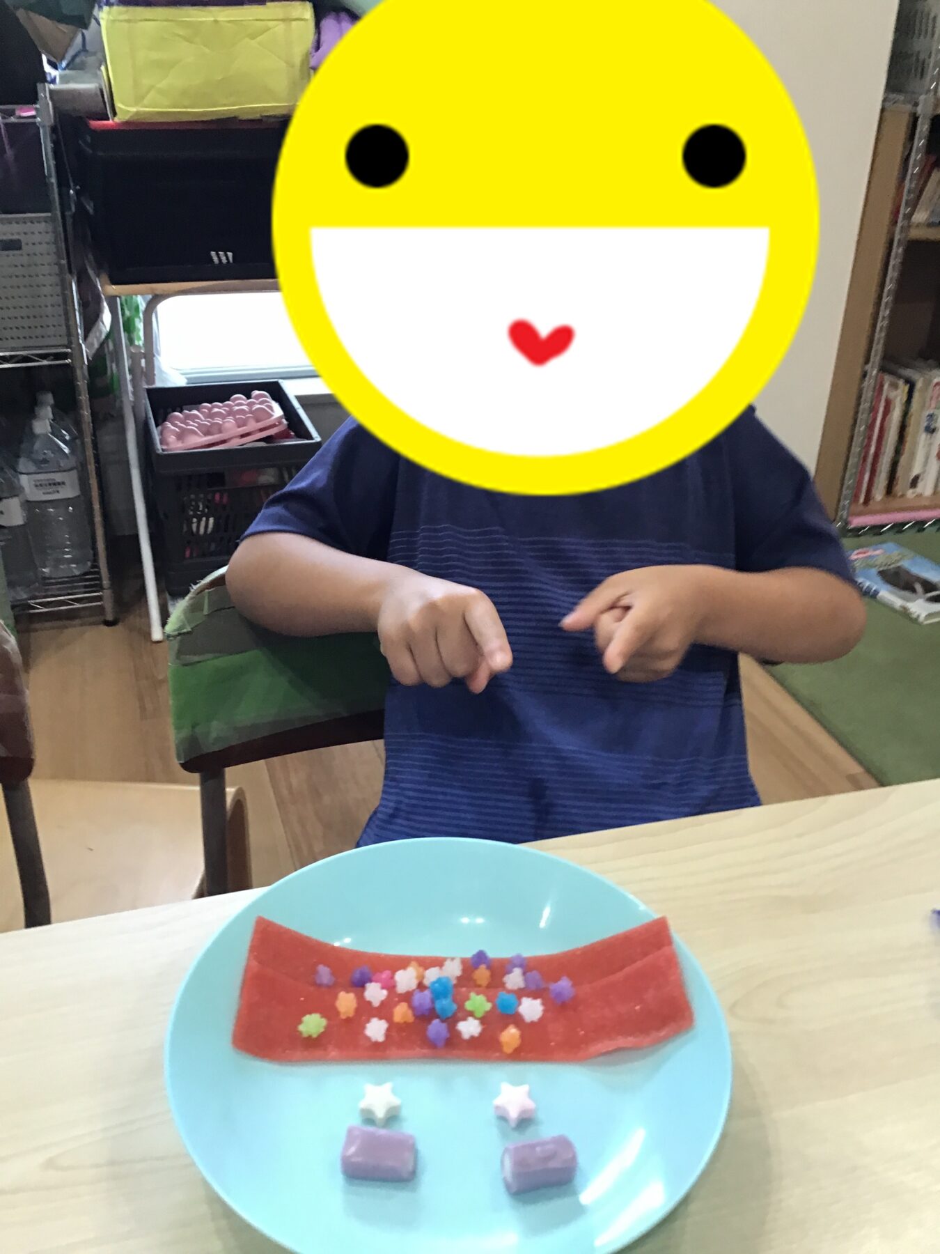 Child at a table with a blue plate holding a red rectangular treat decorated with small colorful candies; the child's face is covered by a large yellow sticker for privacy.