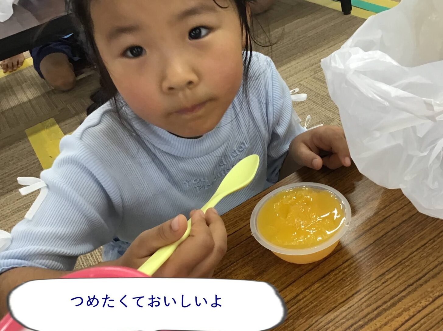 Young child at a table holding a yellow spoon, about to eat orange jelly from a cup, looking at the camera.