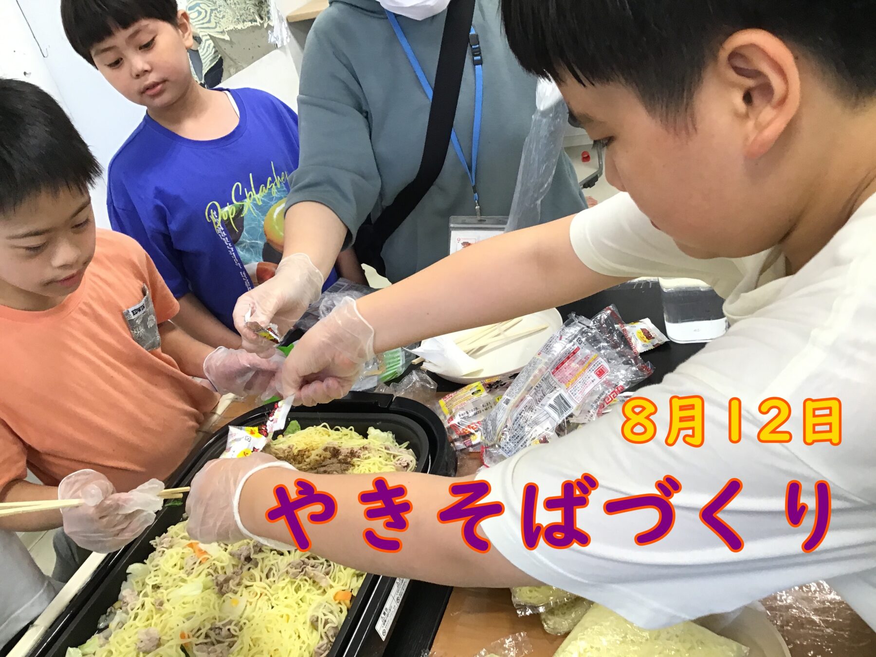 Children wearing gloves assemble and serve yakisoba from a large tray at a table, with noodles and toppings visible and date text overlaid in Japanese