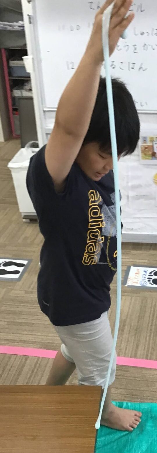 Child barefoot, balancing on a green mat while holding a long blue rope overhead in a classroom.