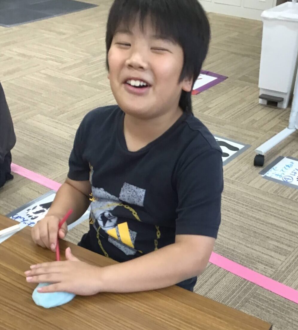 Smiling child sits at a wooden table, shaping light-blue clay with a pink tool in a bright room, pink tape on the floor behind them.
