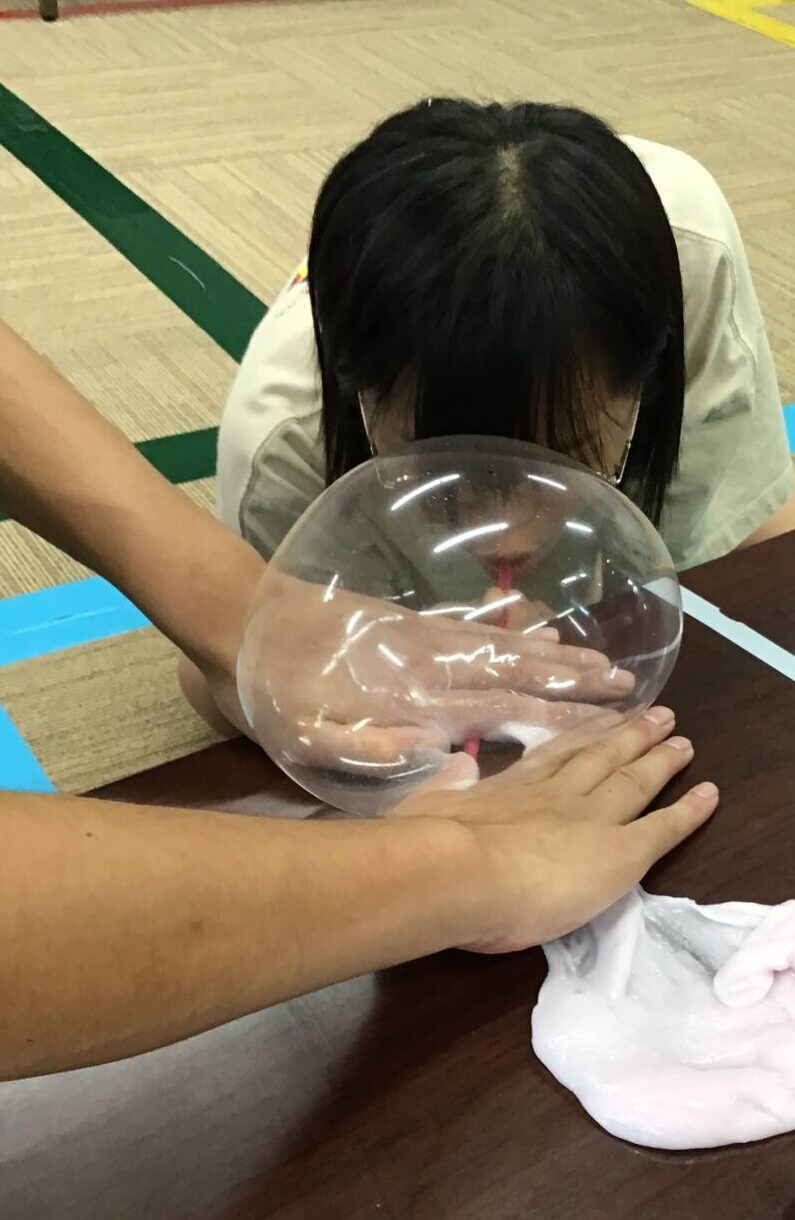 Child seated at a table with head bowed, while an adult presses a large clear balloon against the back of their head. In a gym or classroom setting with colored floor markings in the background.