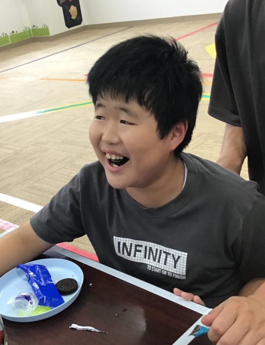 Smiling boy sitting at a table with a red water bottle and blue plate of snacks, while a man stands beside him in a gym-like room.