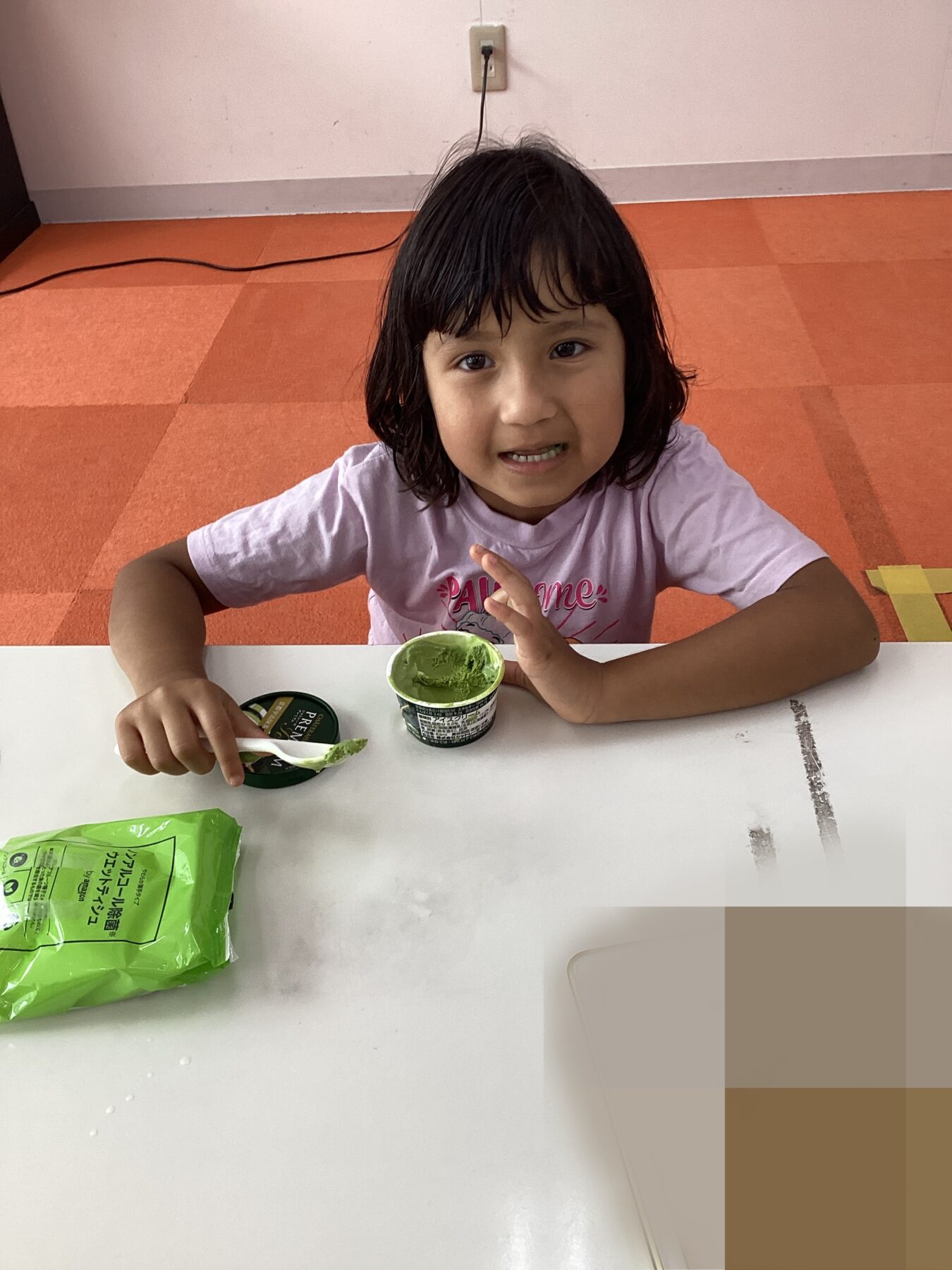 Young child at a white table with a green yogurt cup and spoon, looking at the camera on an orange-tiled floor.