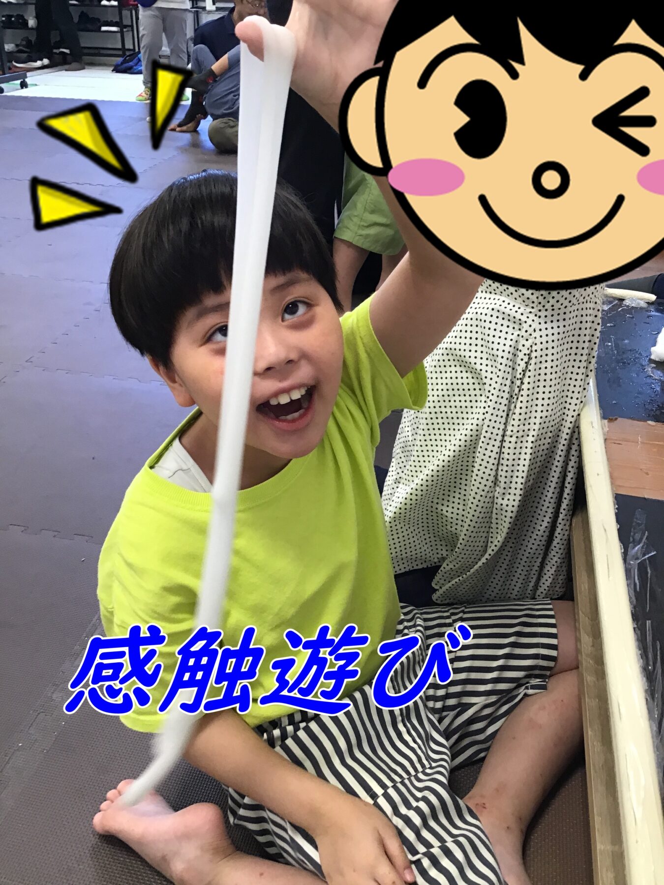 Smiling young boy in a neon yellow shirt holds a white PVC pole overhead in a gym with padded mats and other kids in the background.