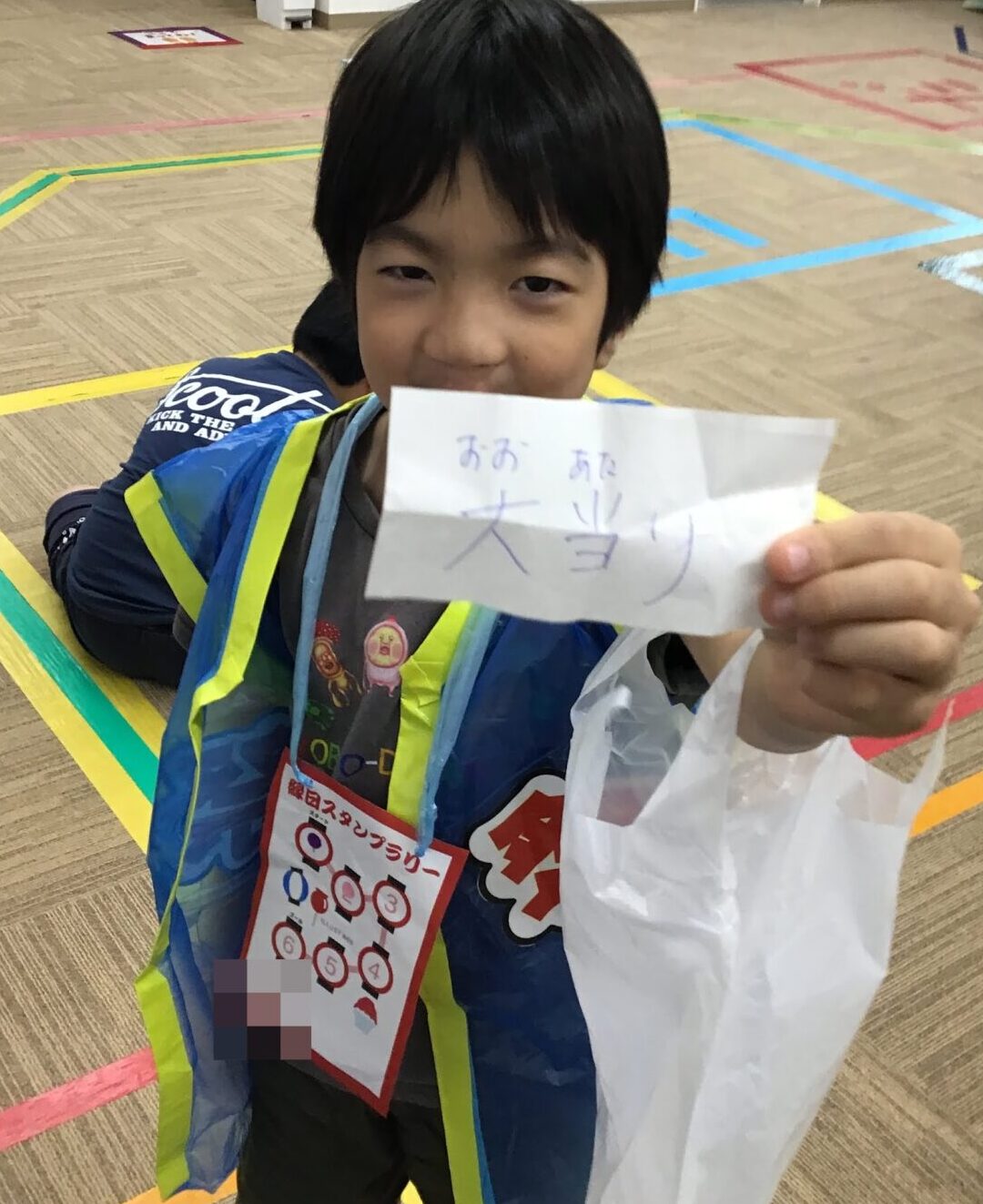 Child in a blue safety vest shows a folded paper with handwritten Japanese text during an indoor event in a room with colored floor markings and a prize bag in hand.