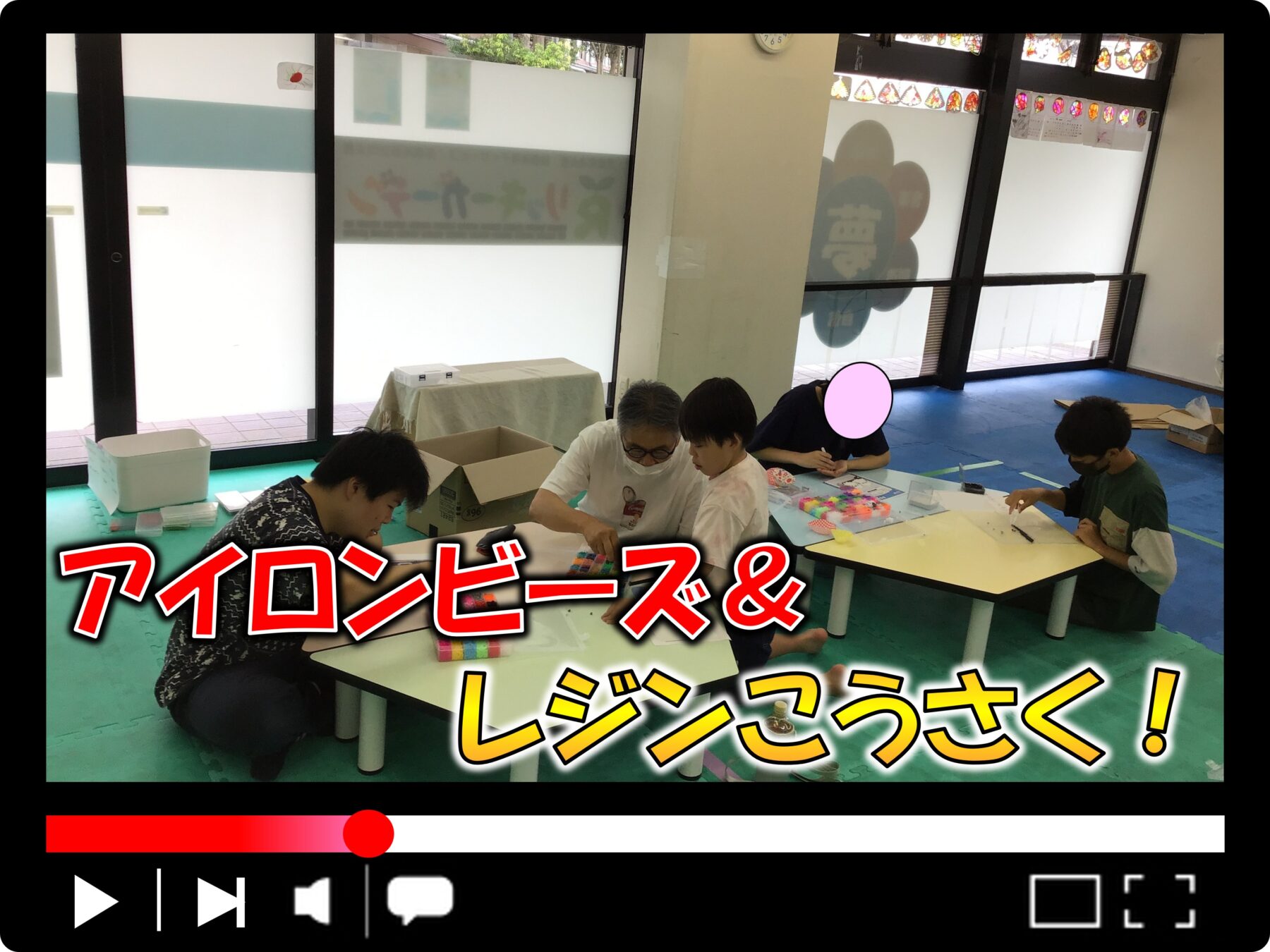 Group of children and an adult crafting at a table in a bright classroom, with art supplies spread out and large windows in the background.