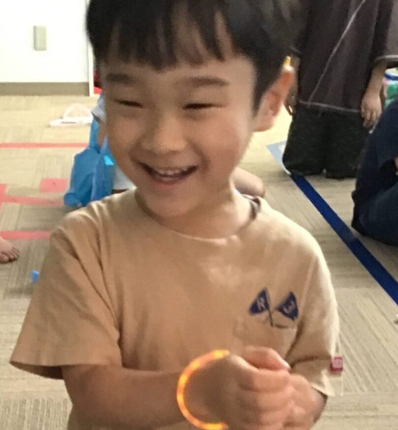 Smiling young boy wearing a light brown shirt and an orange glow bracelet, indoors with others in the background.