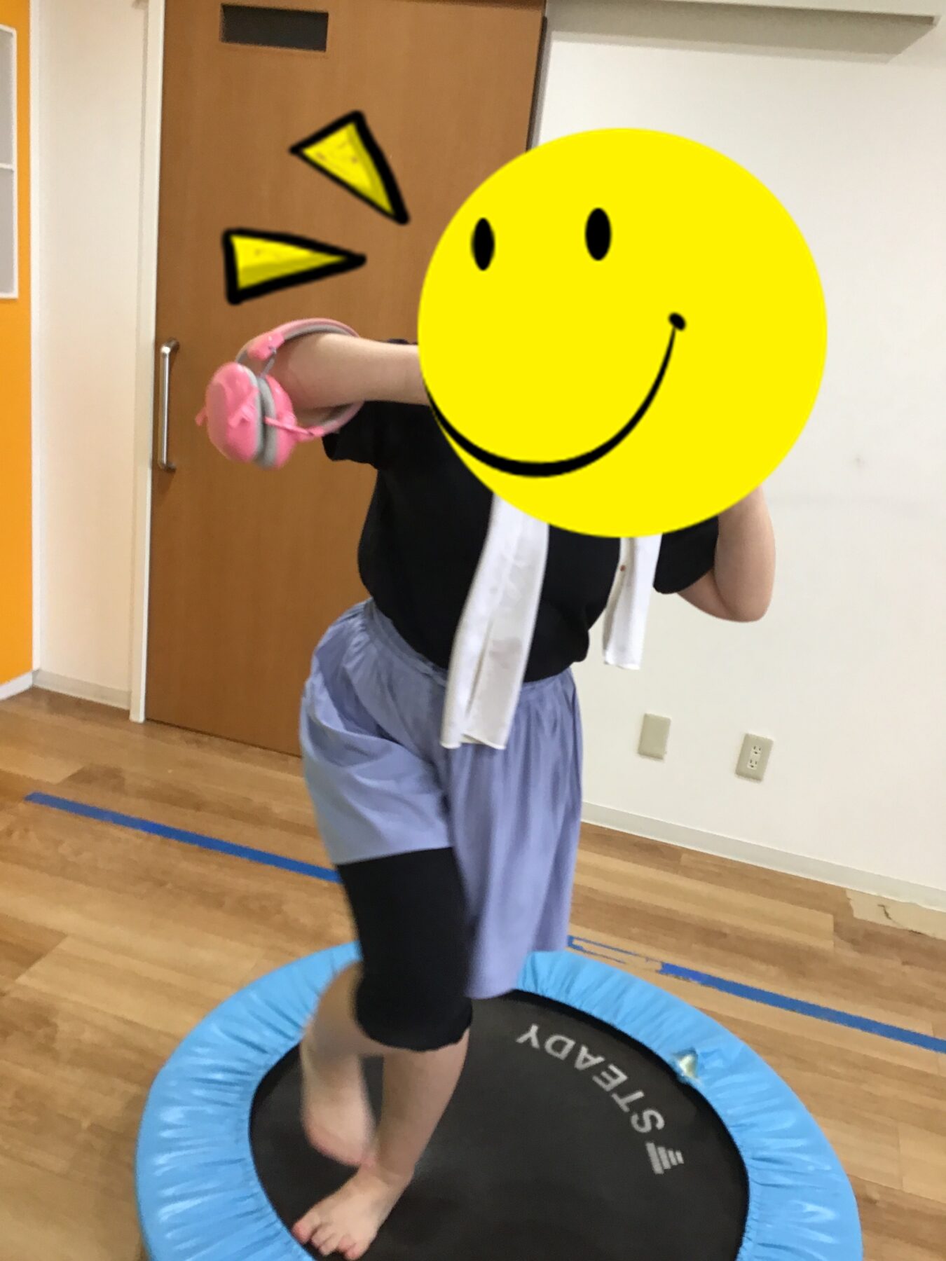 Child standing on a small round trampoline indoors, lifting one leg while wearing a black shirt and light-blue shorts; a large yellow smiley face graphic covers their face.