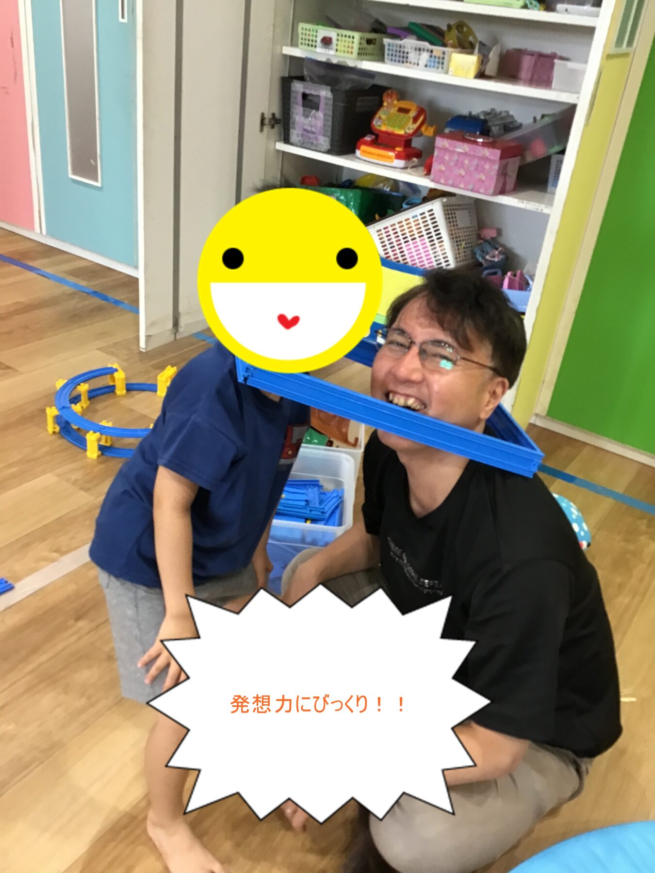 Smiling man and child play with a blue toy train track in a bright playroom; a yellow emoji covers the child’s face and a Japanese caption reads '発想力にびっくり！！'