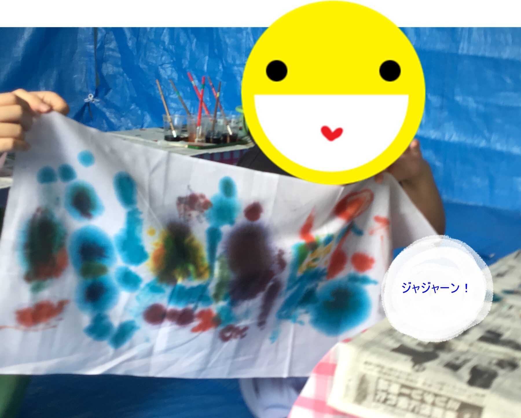 Child holds up a banner smeared with blue and other color handprints against a blue backdrop; yellow smiley face sticker covers their face.