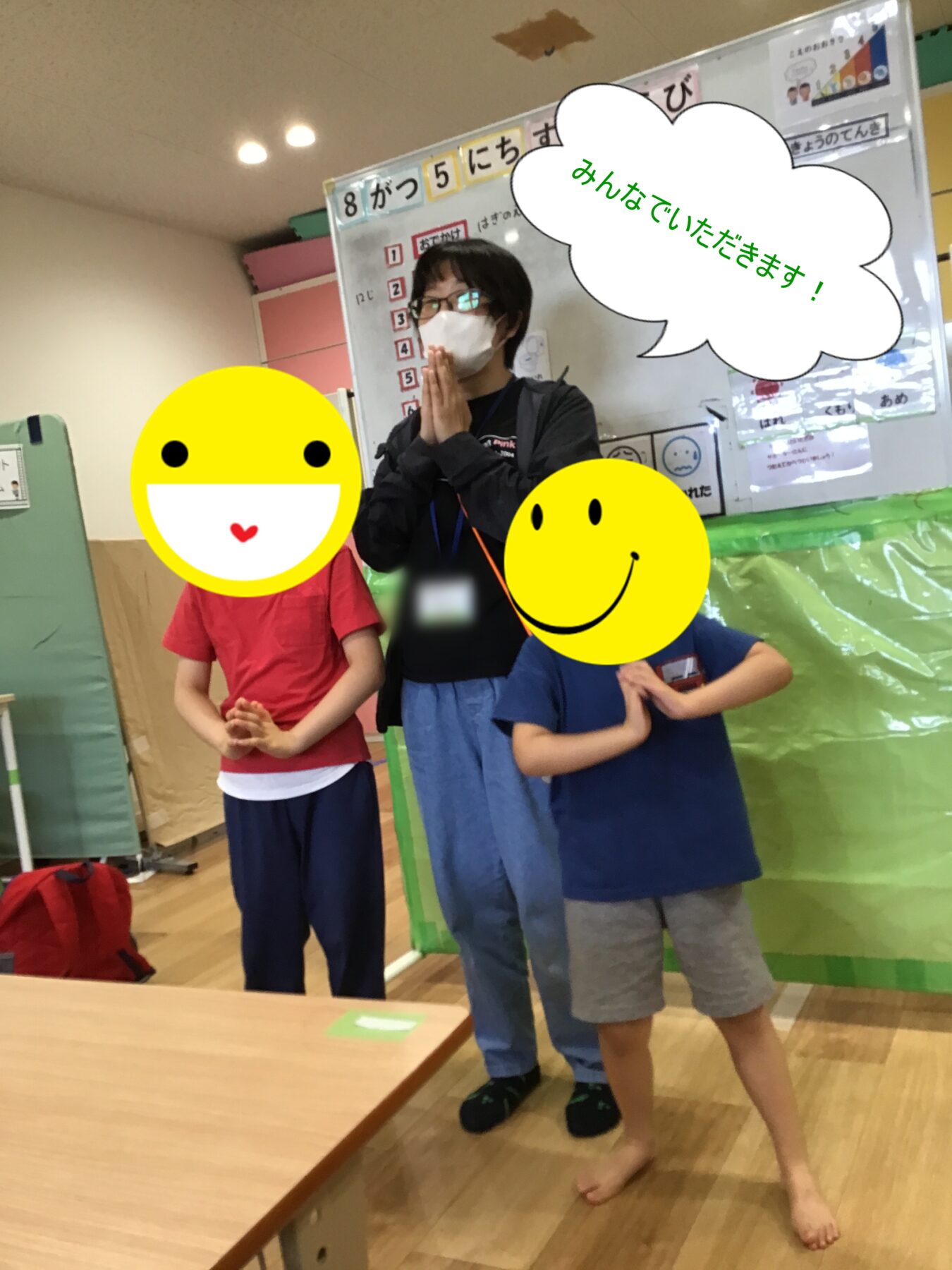 Teacher wearing a mask stands with two children in a classroom, hands pressed in a prayer-like pose as if preparing to eat, with a speech bubble saying 'Let's eat together' in Japanese.