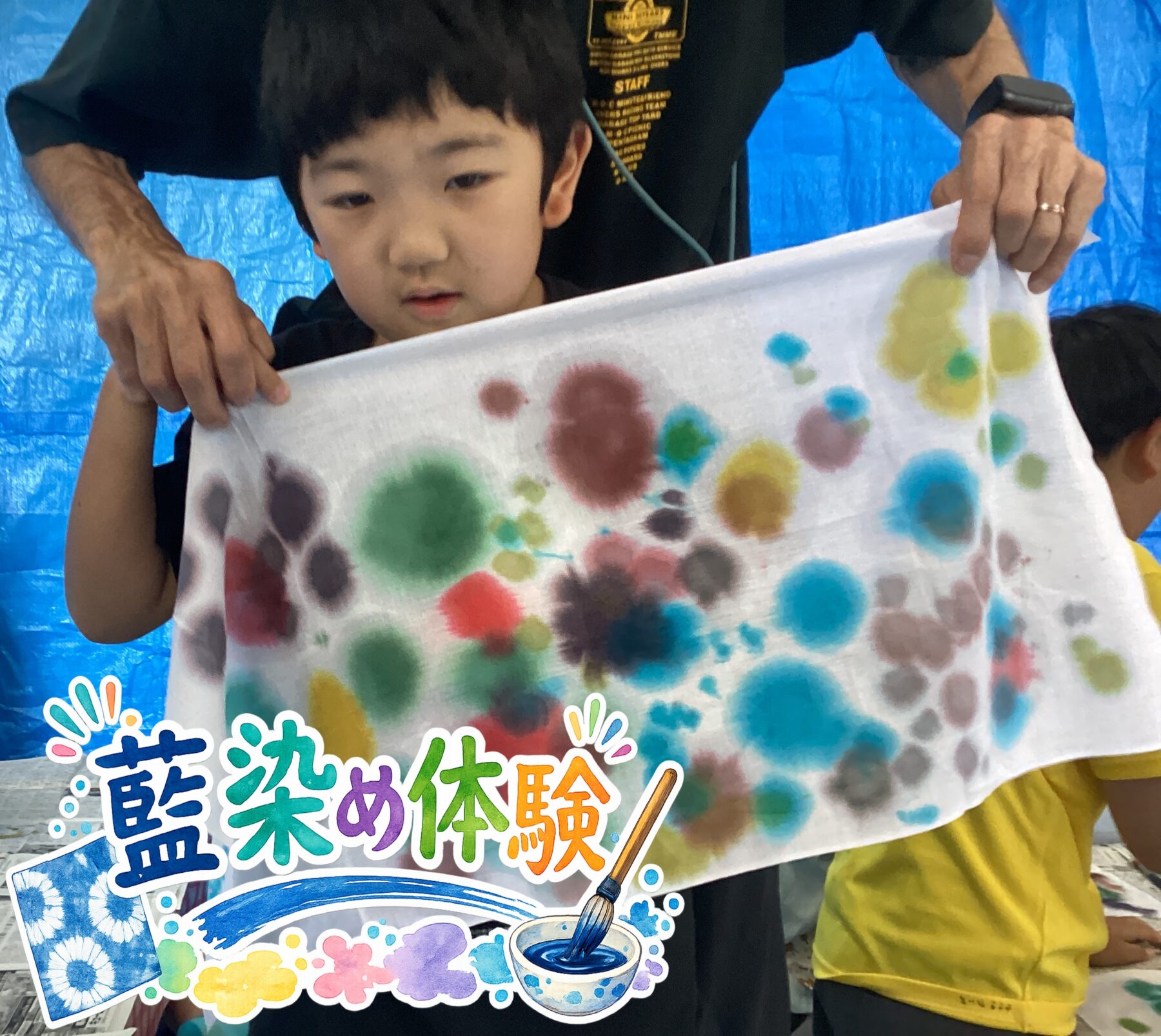 Child holding a white cloth with multicolored paint blobs, assisted by an adult, blue tarp background and colorful sticker at the bottom edge.