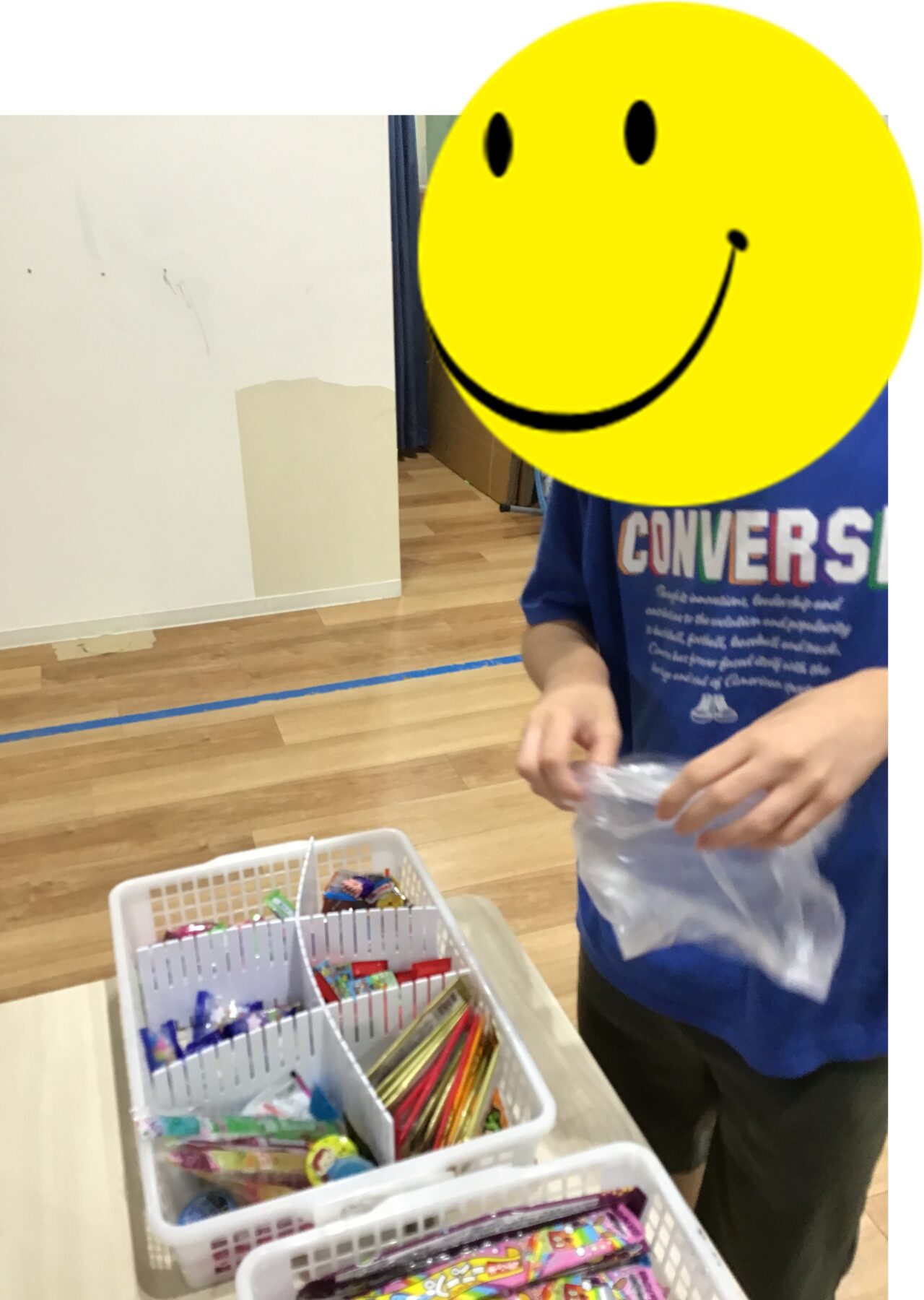Person in a blue shirt sorting colorful candy in white baskets on a wooden gym floor; a large yellow smiley overlay covers their face as a masking sticker.