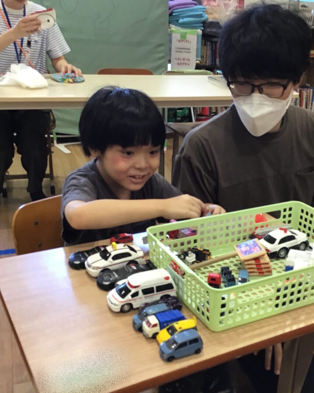 Young boy in a gray shirt sits at a table, playing with toy cars from a green basket, wearing a mask nearby (classroom setting).