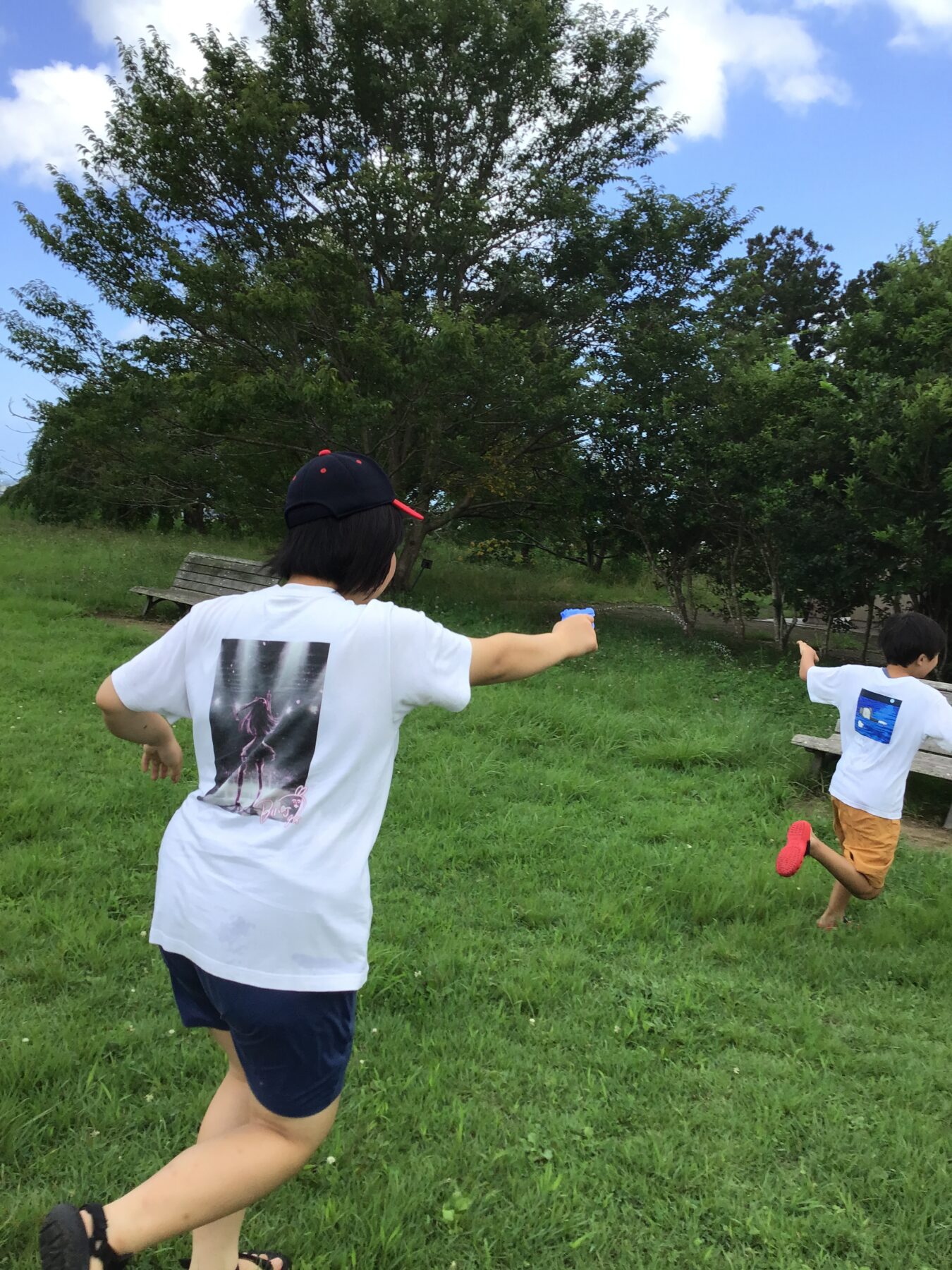Children run across a grassy park, seen from behind, wearing white T-shirts and caps with trees and a bench in the background.