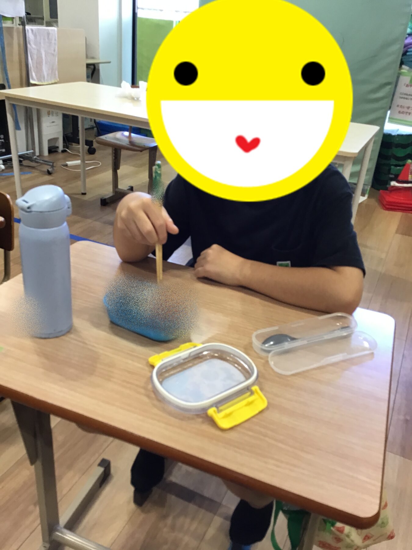 Child seated at a classroom desk, holding a pencil over a small blue oval item, with a light gray water bottle on the table and classroom supplies in the background; face obscured by a large yellow emoji.
