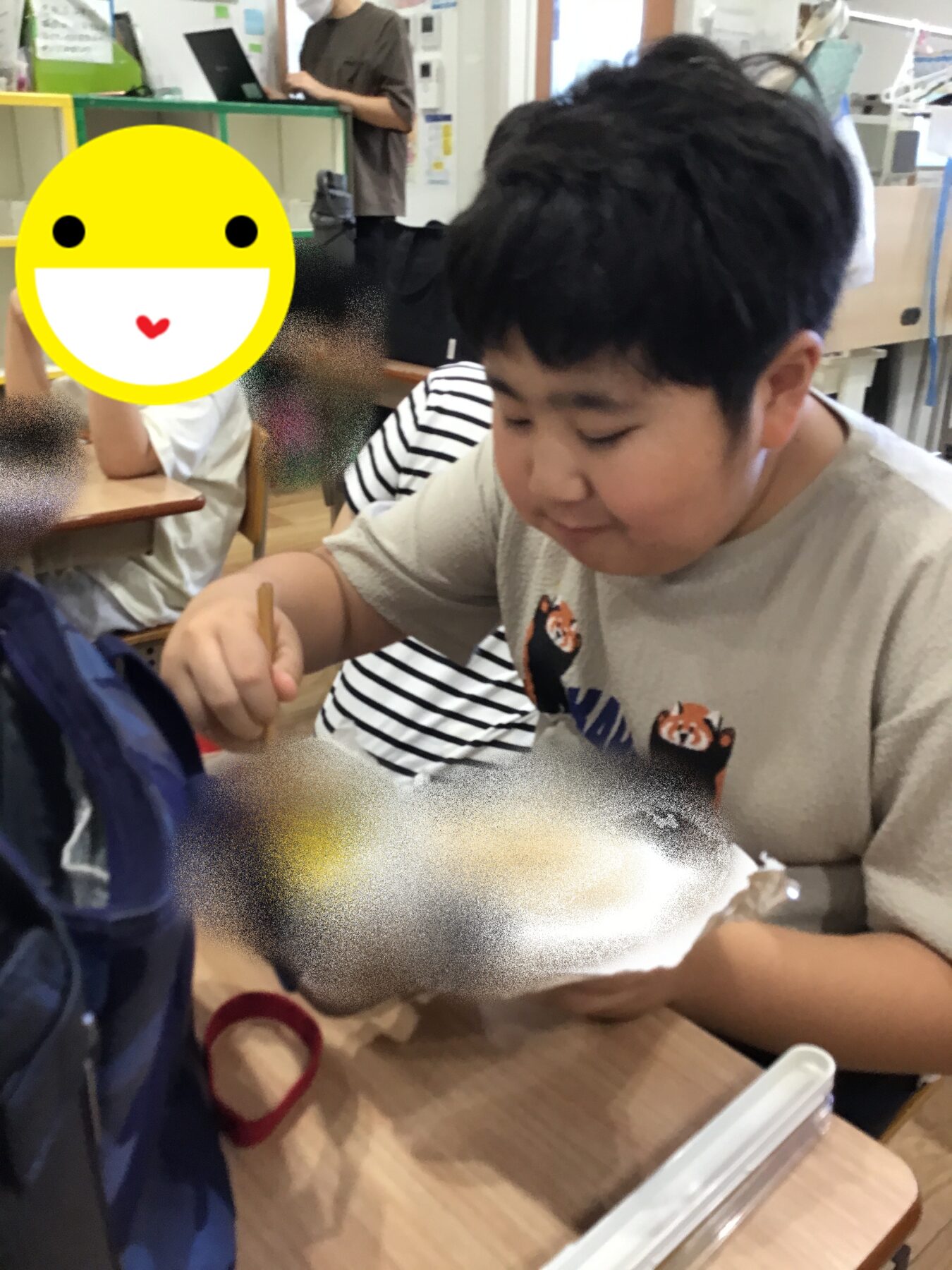Young boy seated at a table, cutting cardboard with scissors, as classmates work nearby in a classroom setting.