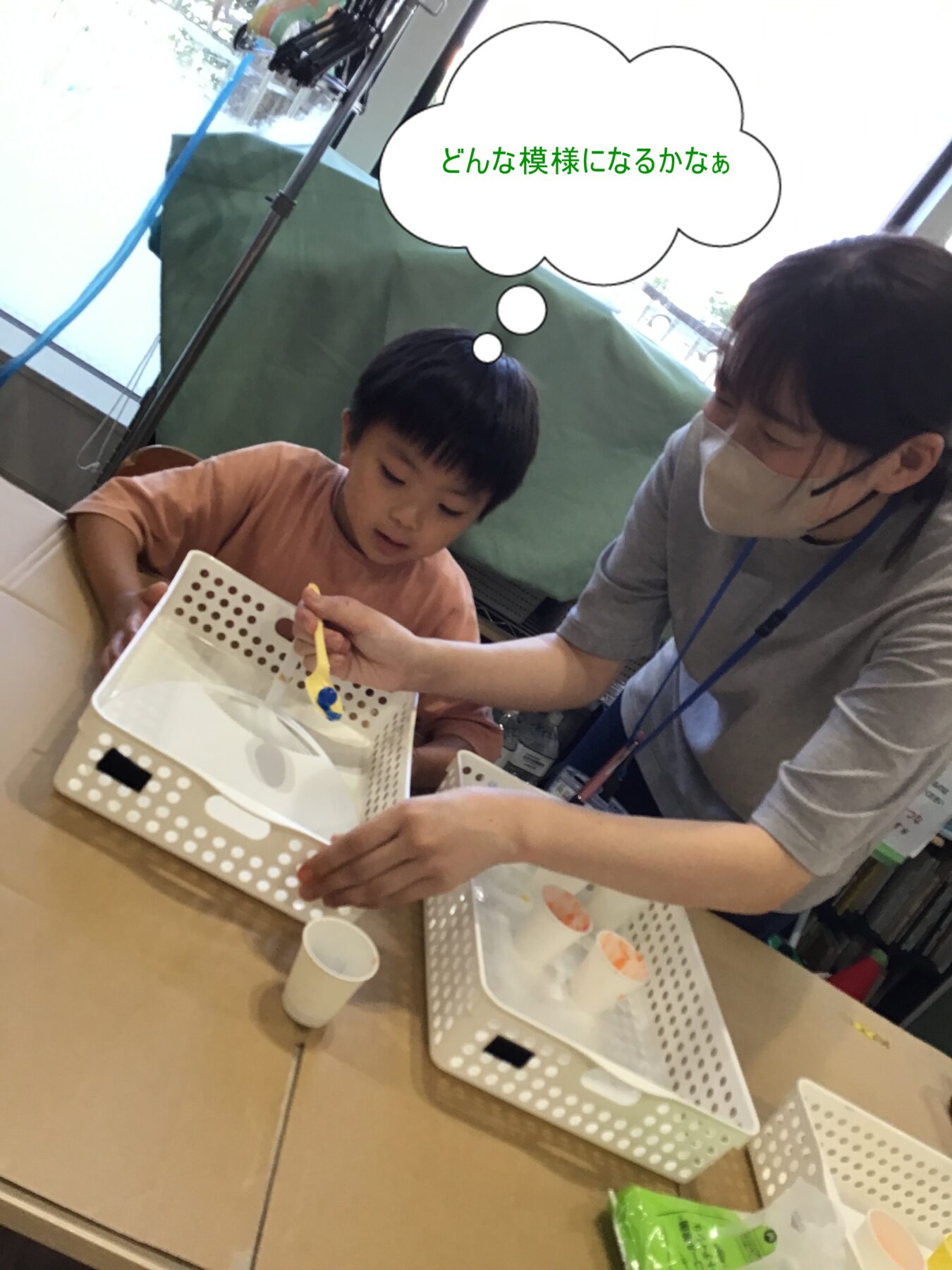 Child and adult wearing masks doing a craft at a table with perforated trays; thought bubble in Japanese asks what pattern will appear.